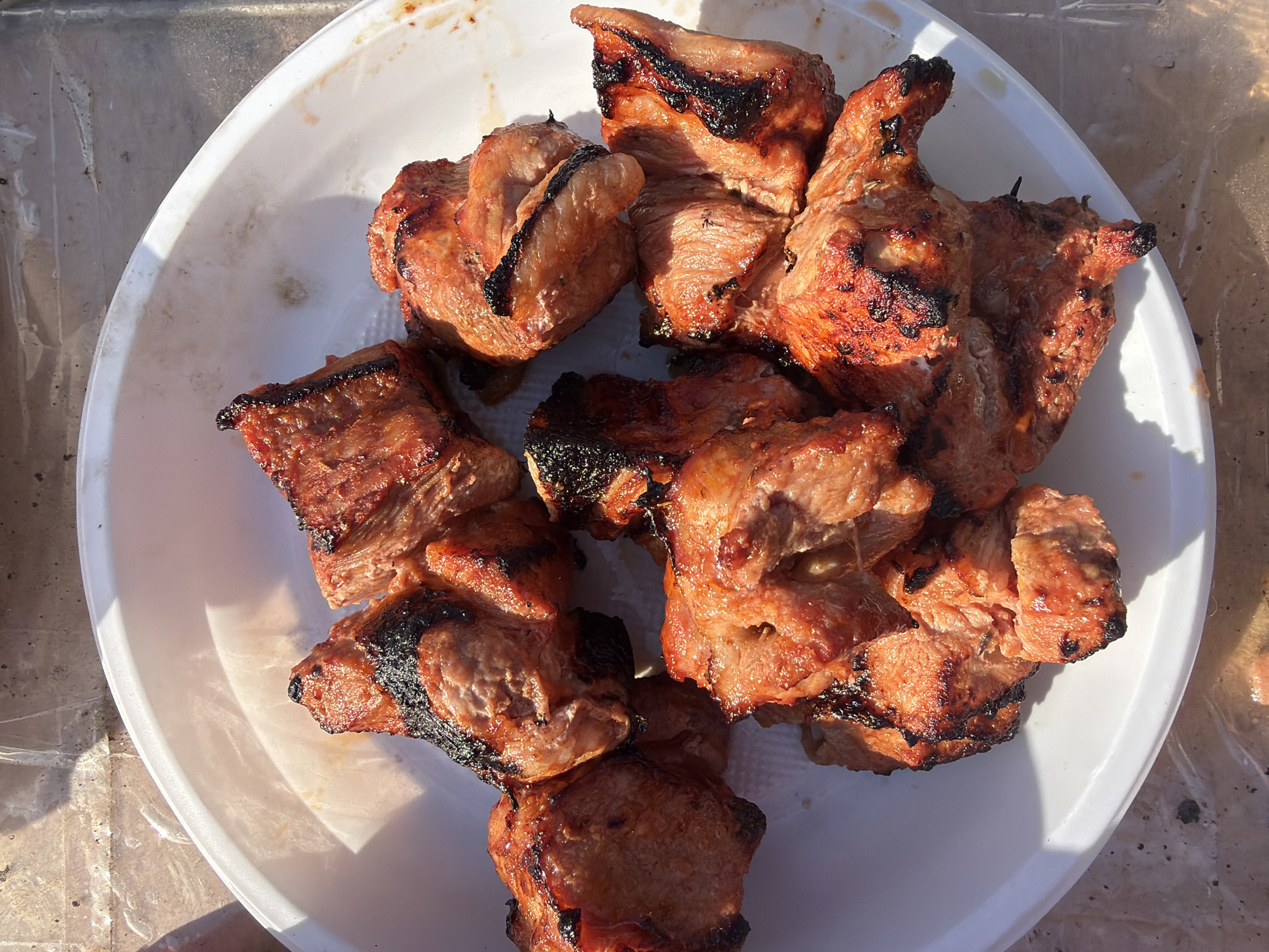 The image shows a white plastic plate filled with pieces of grilled meat. The meat appears to be pork due to its color and texture, and it has visible grill marks. The plate is placed outdoors, possibly on a table, judging by the blurred background and lighting. The lighting suggests it is daytime. The image does not reveal any specific details that would help identify it as being located in Chaikovsky, Russia. There are no people or other objects in the frame.