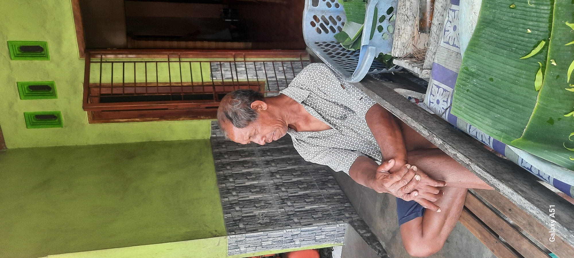 An elderly man with graying hair and fair skin is sitting on a wooden bench outdoors. He is wearing a patterned short-sleeved shirt and dark blue shorts. His hands are clasped in his lap. To his right, a blue plastic basket contains sliced banana leaves, and a wooden crate is visible. The background features a light green wall with decorative grey and white tiles. A wooden door with metal bars is slightly ajar, revealing an interior space. The scene appears to be in Selopuro, Indonesia, as indicated by the visible text "Galaxy A51" on the lower left corner of the image. The overall impression is a snapshot of daily life in a rural or suburban Indonesian setting.