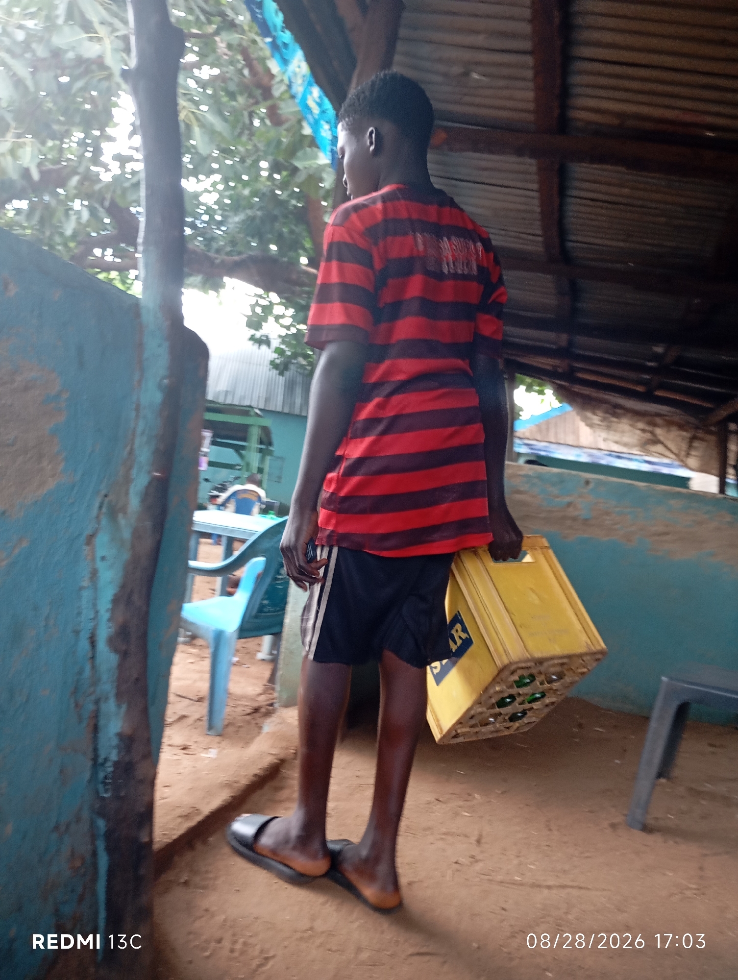 In Shendam, Nigeria, a young Black male wearing a red and black striped t-shirt and black shorts carries a yellow crate of bottled beverages. He is walking away from the camera, towards a structure with a corrugated metal roof supported by wooden beams. To his left, a blue wall partially obscures the view of tables and chairs in an outdoor area. The ground is sandy. The timestamp "08/28/2026 17:03" and "REDMI 13C" are visible in the lower portion of the image.