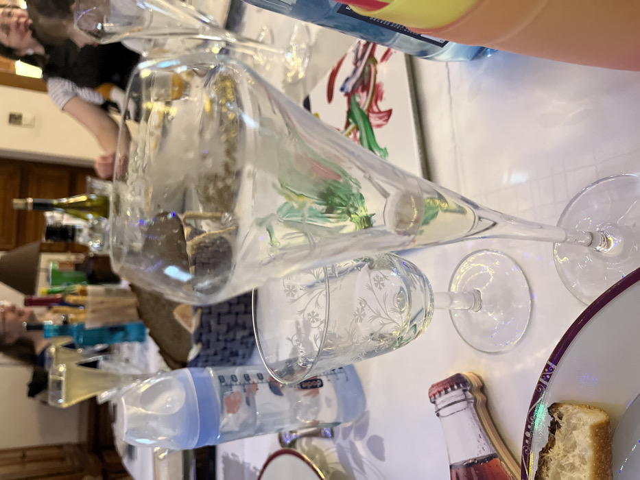 The image depicts an indoor dining table scene within a residence in Ancenis, France. The table is covered with a white tablecloth. In the foreground, a white plate with a maroon border holds a piece of bread. To its right, a clear stemmed glass with etched floral patterns stands beside a tall, clear champagne flute containing a pale yellow liquid and reflecting ambient colors. A light blue plastic baby bottle, marked "Chicco," filled with clear liquid, is positioned behind these glasses. A flat, bottle-shaped decorative object with a red cap rests on the tablecloth. A stack of colorful (yellow, orange, pink) plastic cups is partially visible on the far right.
In the midground, a dark blue woven basket holds multiple slices of dark bread. Various bottles, including a prominent blue bottle and a green bottle, are interspersed among other glasses and champagne flutes, some containing a pale yellow beverage. A white square plate with abstract green, red, and blue paint strokes is visible behind the champagne flute.
The background, out of focus, shows several individuals seated around the table. A woman with dark hair and glasses is visible on the left, and another woman with dark hair, glasses, wearing a striped top and a dark vest, is seen smiling on the right. A table lamp with a dark shade is centrally located in the blurred background, with a wooden furniture piece visible on the far left. The scene suggests a social gathering or meal.