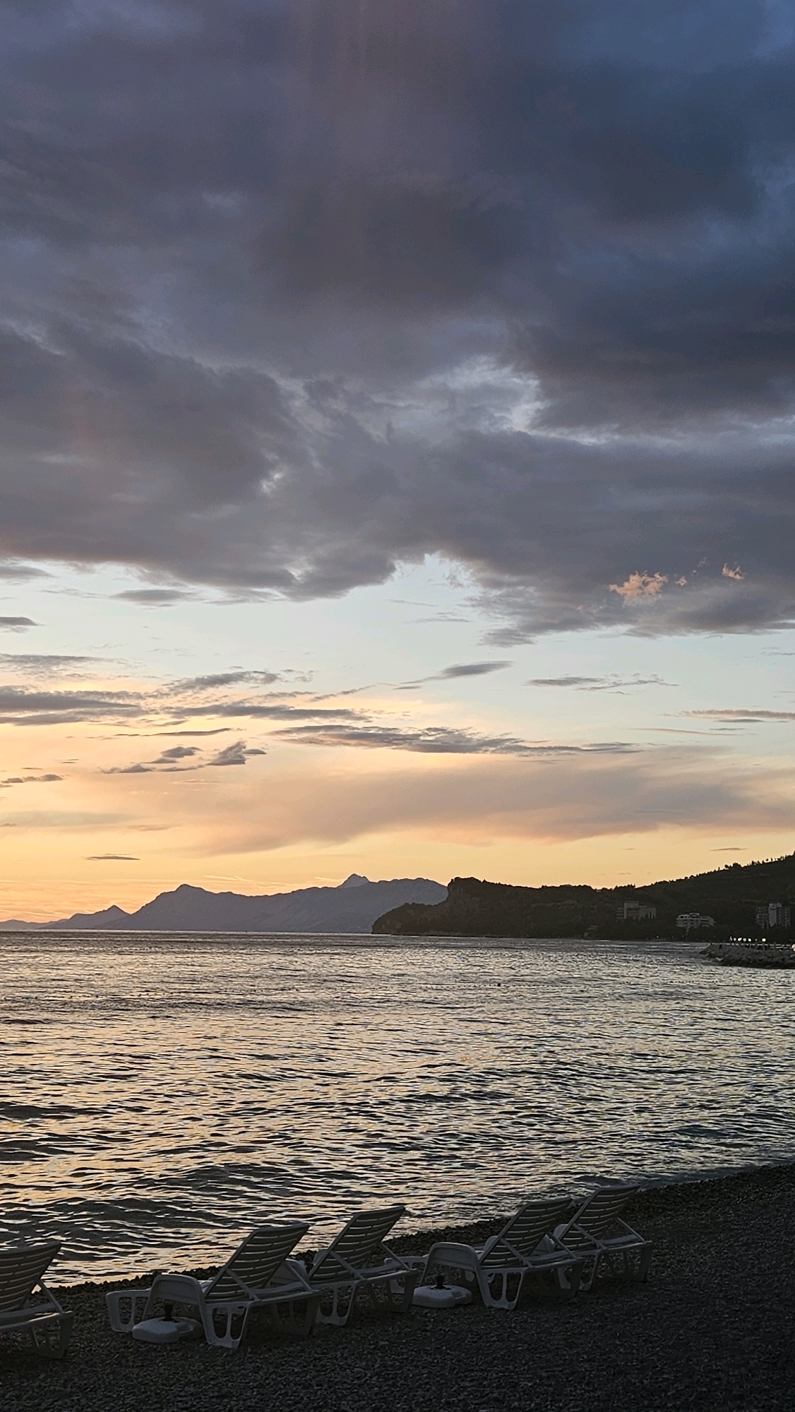 The image depicts a serene beach scene at dusk in Općina Tučepi, Croatia. In the foreground, several empty lounge chairs are arranged on a pebble beach, facing the calm sea. The water reflects the soft hues of the sunset. In the middle ground, a distant coastline is visible, with rolling hills and a few buildings nestled along the shore. The background is dominated by a dramatic sky filled with dark, moody clouds interspersed with patches of pale orange and pink light from the setting sun. The overall impression is one of tranquility and natural beauty, with the natural elements of the sea, sky, and land creating a picturesque landscape.