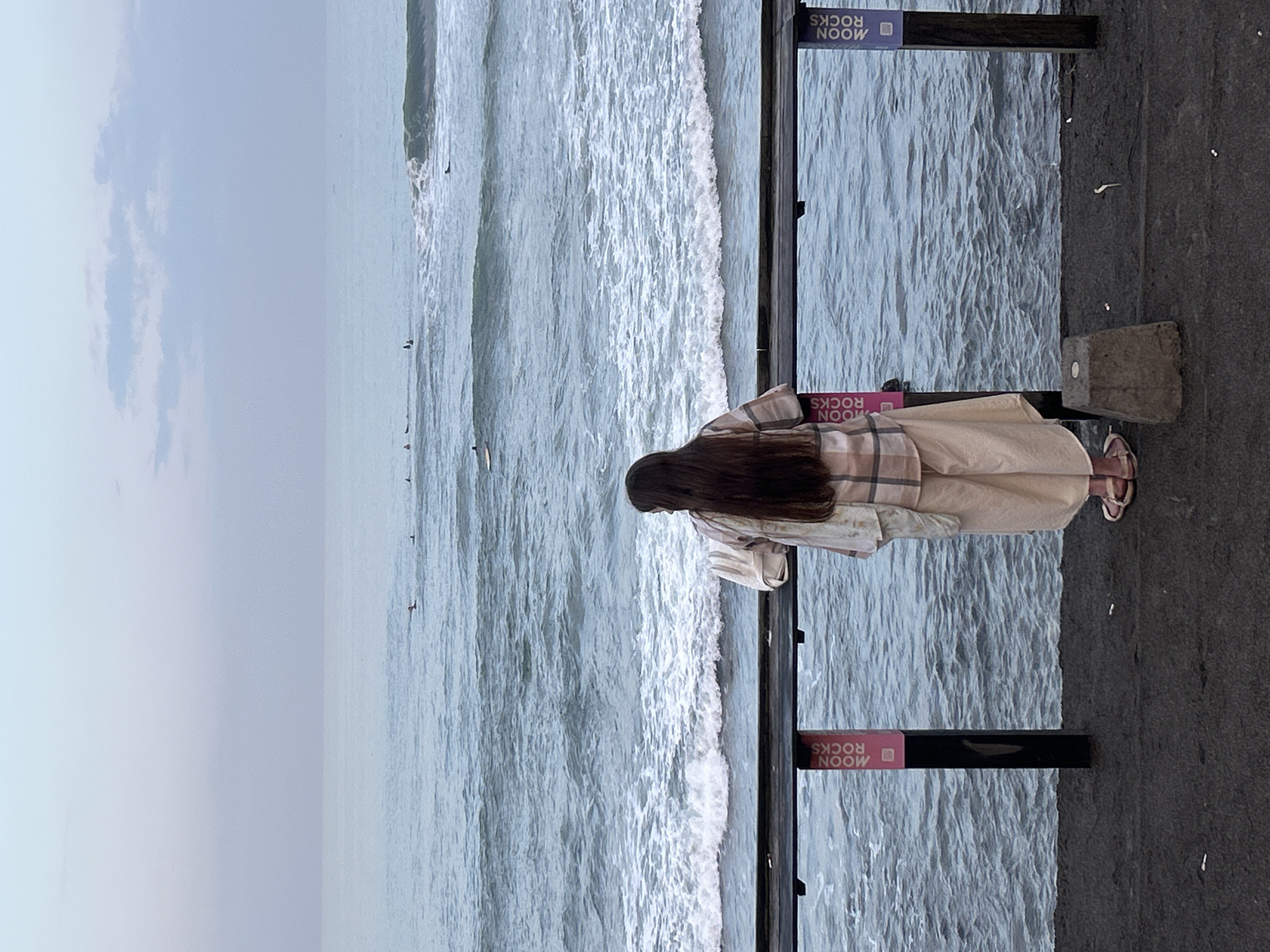 An outdoor, eye-level, medium shot shows a person from the back, facing the ocean from a railing. The person, with long, dark brown hair, wears a plaid shirt and light-colored, loose-fitting pants. Their feet are in light-colored sandals. They are leaning on a dark railing, which has two vertical signs attached to it. The sign on the left is pink and reads "MOON ROCKS" in white text, with a small square icon below. The sign on the right is purple and reads "MOON ROCKS" in white text. The ocean is visible behind the railing, with waves breaking and several surfers in the water. The sky is overcast with pale blue and gray tones. The ground in front of the railing is dark and appears to be asphalt or concrete, with a small, light gray concrete block in the foreground. The scene appears to be in North Kuta, Indonesia, likely near a beach or a coastal promenade.