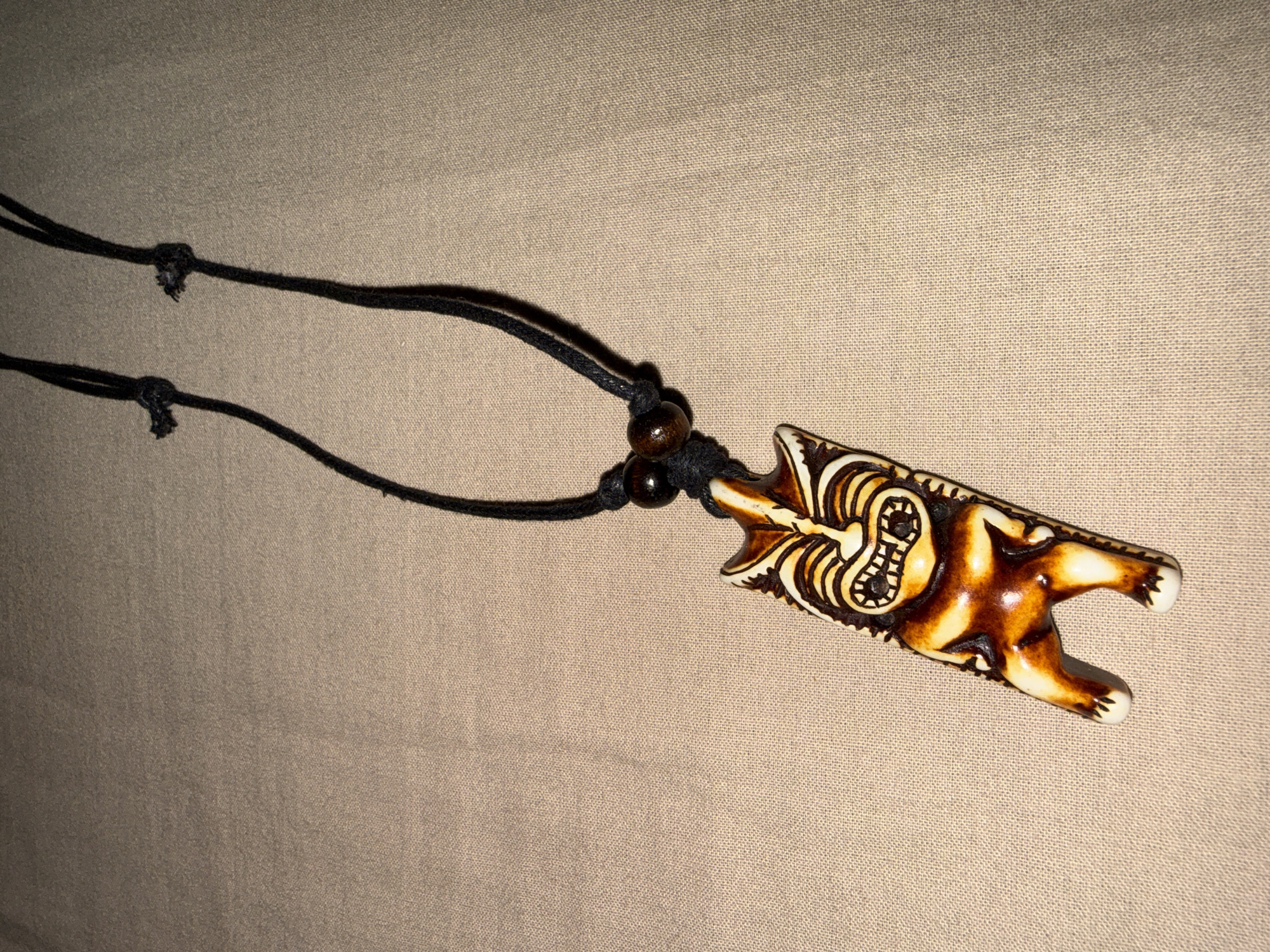 A close-up, top-down shot shows a tiki pendant necklace lying on a beige, textured fabric surface. The pendant is carved from a light-colored material with dark brown accents, depicting a stylized tiki figure with a wide grin and protruding eyes. The pendant is suspended from a black cord, which is knotted at the top, with two dark wooden beads on either side of the pendant. The background is a uniform beige fabric. There is no indication of the city of Tsarevo, Bulgaria.