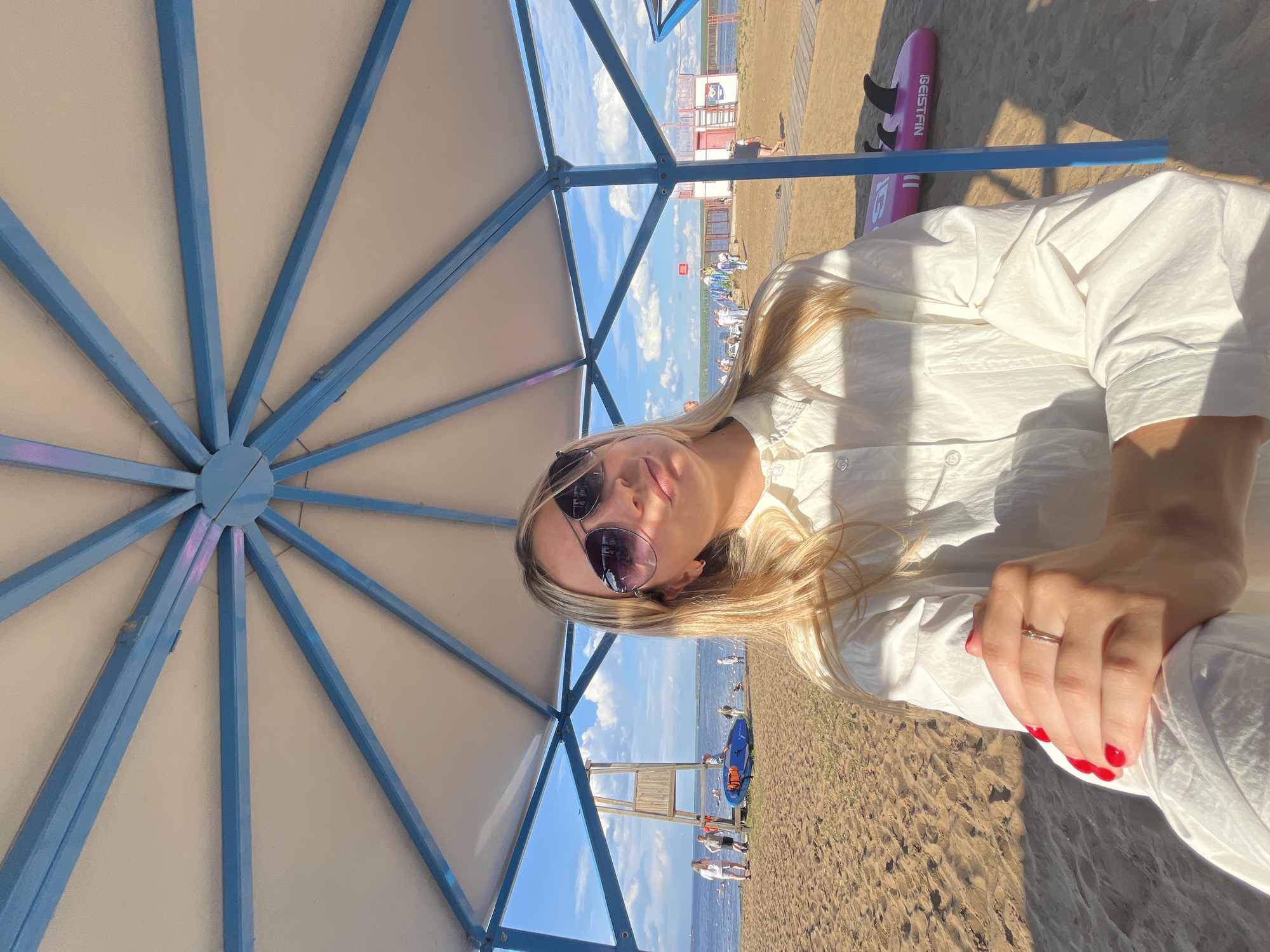 A young woman with blonde hair and sunglasses is taking a selfie on a sandy beach under a light-colored umbrella with a blue frame. She is wearing a white button-down shirt. Her right hand, with red nail polish and a ring, rests on her lap. In the background, there is a body of water, a lifeguard tower, several people, and a blue paddleboard with an orange life vest. A pink surfboard with the text "GEISTFIN" is visible to the right of the woman. The sky is blue with scattered white clouds, indicating a sunny day. The scene appears to be during daylight hours. The overall mood is relaxed and summery.