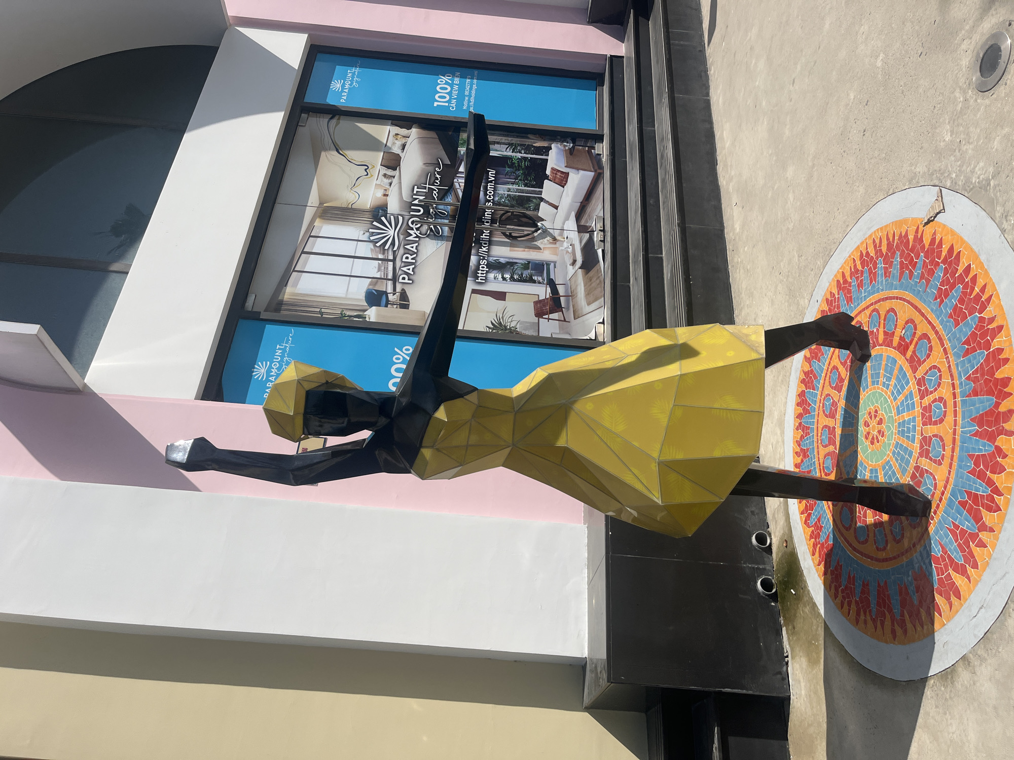 A black and yellow geometric sculpture of a woman in a dress stands on a circular mosaic platform. The sculpture is positioned in front of a building with a large glass window displaying advertisements for "PARAMOUNT Signature". The building has a pale yellow, white, and pink facade. The scene appears to be an outdoor public space, possibly a commercial area in Nha Trang, Vietnam, given the Vietnamese text on the advertisement. The lighting suggests it is daytime, with shadows cast by the sculpture and building.