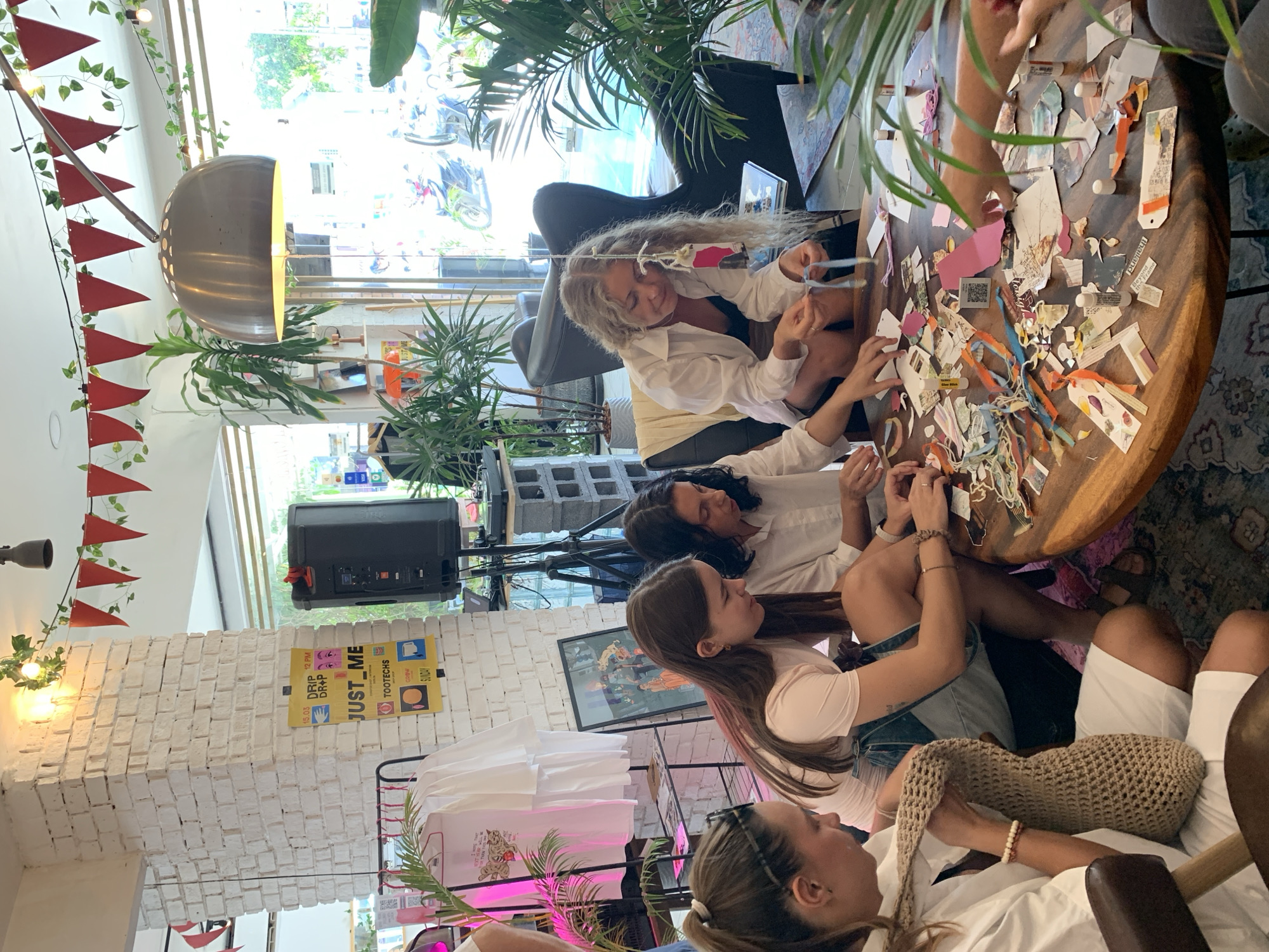 The image depicts an indoor gathering of at least four individuals engaged in a crafting activity around a circular wooden table laden with paper scraps, scissors, glue sticks, and various decorative elements. The setting appears to be a bright, airy space with large windows that offer a view of an outdoor scene featuring motorcycles and lush greenery, suggesting a tropical location. Red triangular flags and greenery are strung across the ceiling, adding a festive atmosphere. To the left, a white brick wall is visible with a framed poster and clothing items displayed on a rack. The individuals, appearing to be women of varying ages, are focused on their creative task, with some interacting with the materials and others looking on. The overall impression is that of a collaborative workshop or social event.