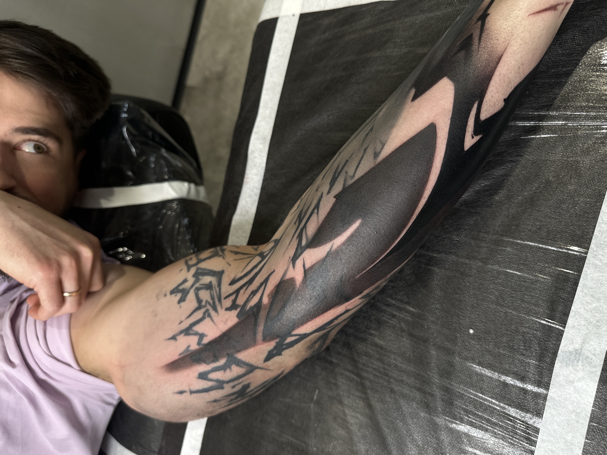 A high-angle, medium shot captures a person's arm adorned with a black abstract tattoo. The tattoo covers a significant portion of the forearm and extends towards the upper arm. The design incorporates sharp, angular shapes with negative space, giving it a graphic and modern aesthetic. The person is wearing a light purple t-shirt and has a ring on one of their fingers. The individual is partially visible, showing their head and eye, hinting at fair skin and brown hair. The arm rests on a black surface, possibly a tattoo chair or bed, with a white stripe marking its edge. The background is slightly blurred, but it is suggestive of a clinical or studio-like environment. I don't have the information about this image location in Moscow, Russia.