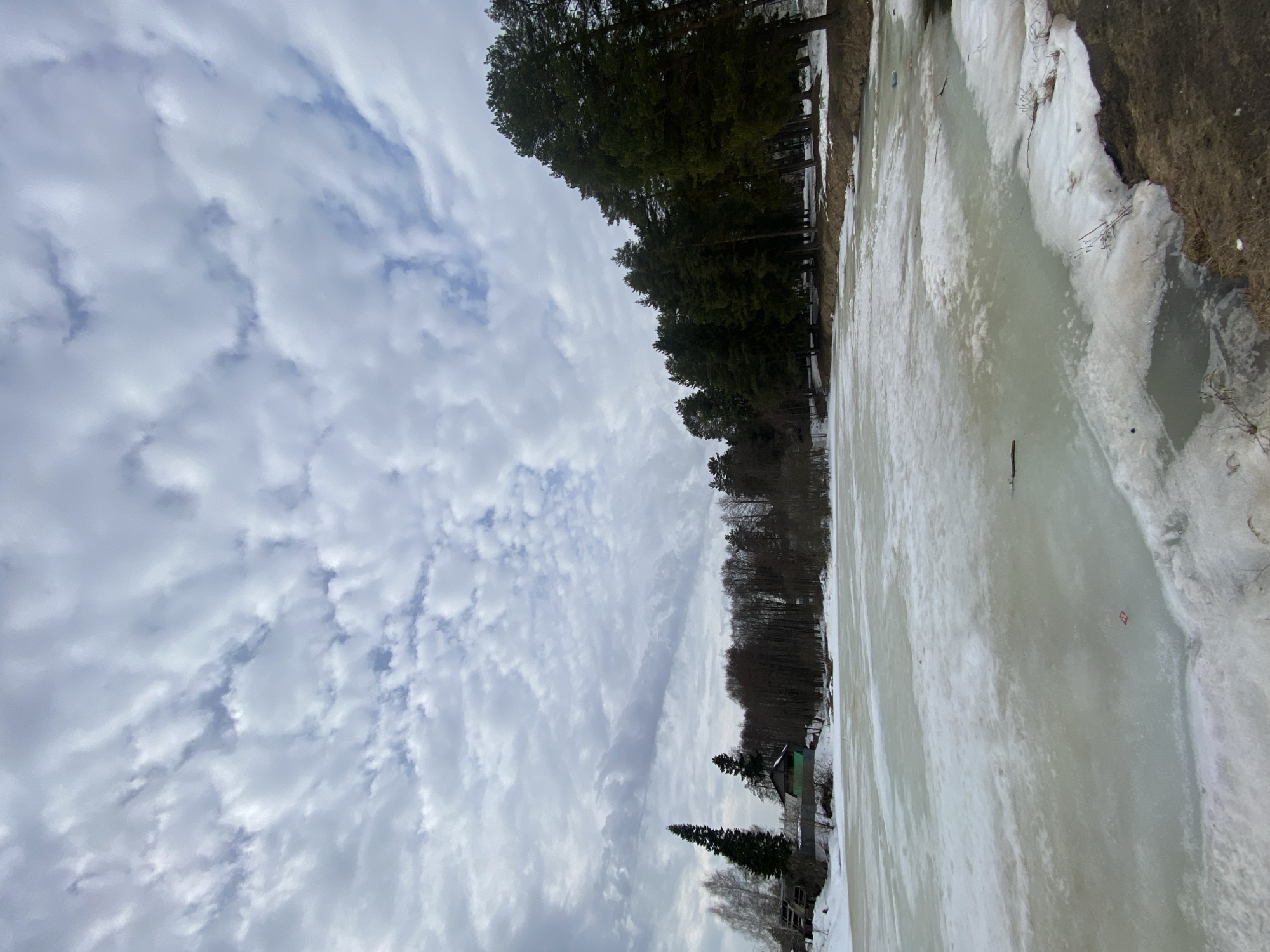 The image depicts a partially frozen body of water in what appears to be a rural or semi-rural location near the city of Chaikovsky, Russia. The foreground shows a frozen lake with sections of ice and patches of melting snow along the edges. A shoreline is visible, characterized by brown vegetation and trees. Coniferous trees line the distant shore. In the mid-ground, there's a small structure, possibly a house or shed, nestled among the trees. Above, the sky is filled with dense, layered clouds, indicating overcast weather. The scene gives the impression of early spring, where winter's hold is gradually weakening.