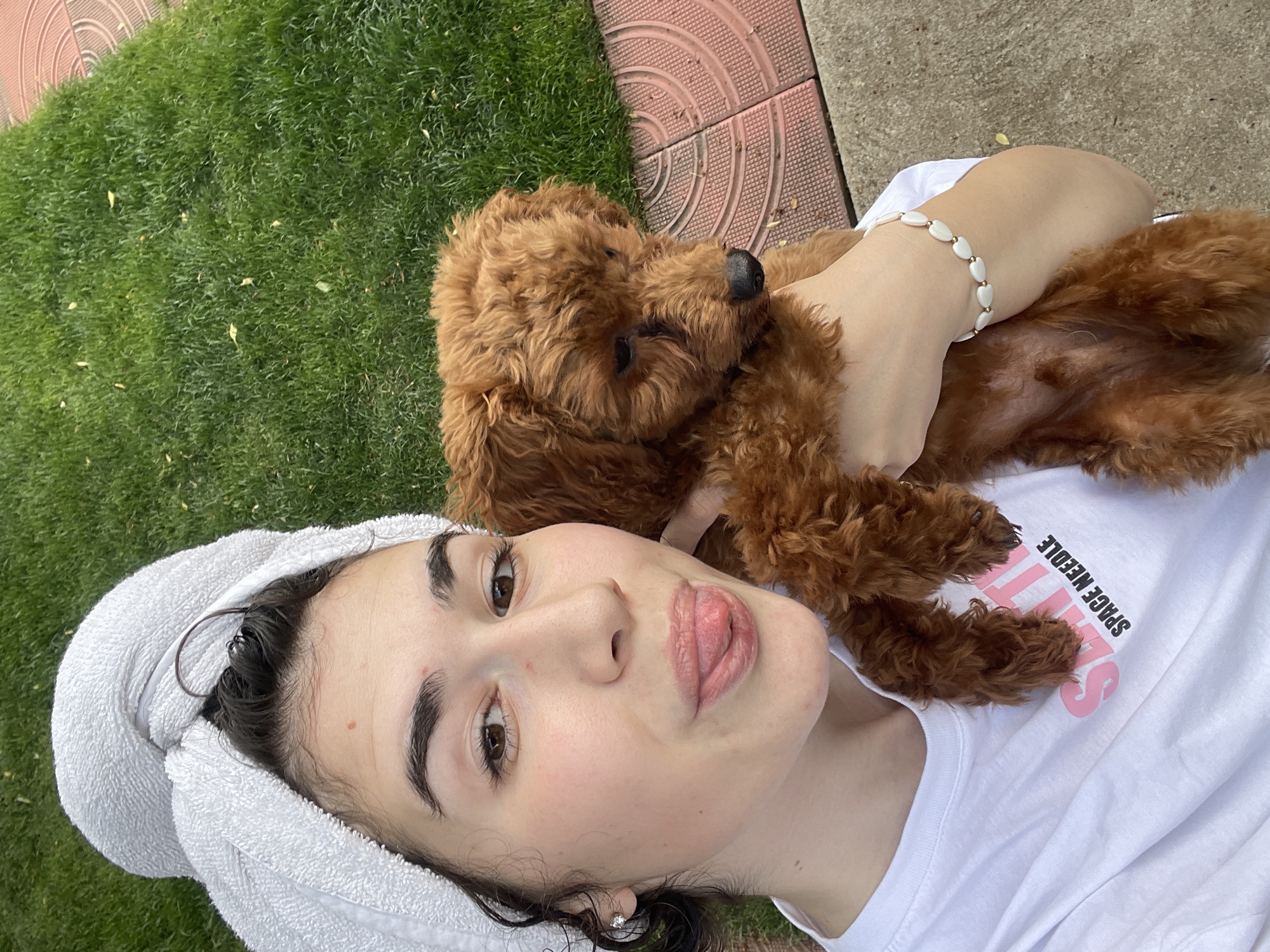A young woman with a white towel wrapped around her head lies on her back on the grass next to a tiled walkway. She wears a white t-shirt that reads "Seattle Space Needle" in pink lettering. A small brown poodle lies on her chest, cradled by her hand. The location cannot be determined as Sofia, Bulgaria, from the image alone.