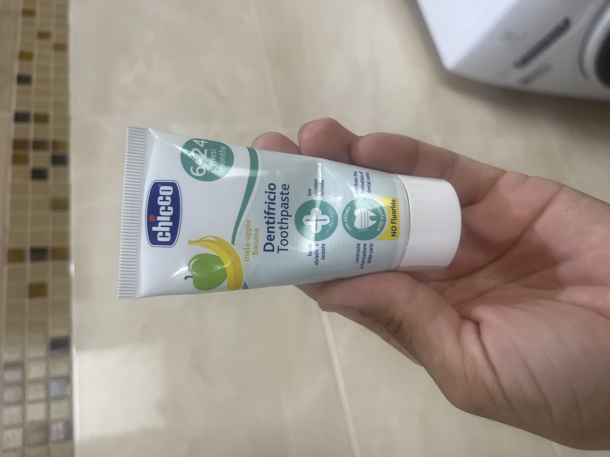 A close-up shot shows a hand holding a tube of Chicco baby toothpaste. The toothpaste tube is white with green and blue lettering. The flavor is described as "mela-apple banana". The tube also indicates that it is suitable for babies aged 6-24 months and has a "low abrasion formula" and is "with xylitol." A prominent label states "NO Fluoride". The background appears to be a tiled wall, possibly in a bathroom. The image was captured in Onokivtsi, Ukraine.