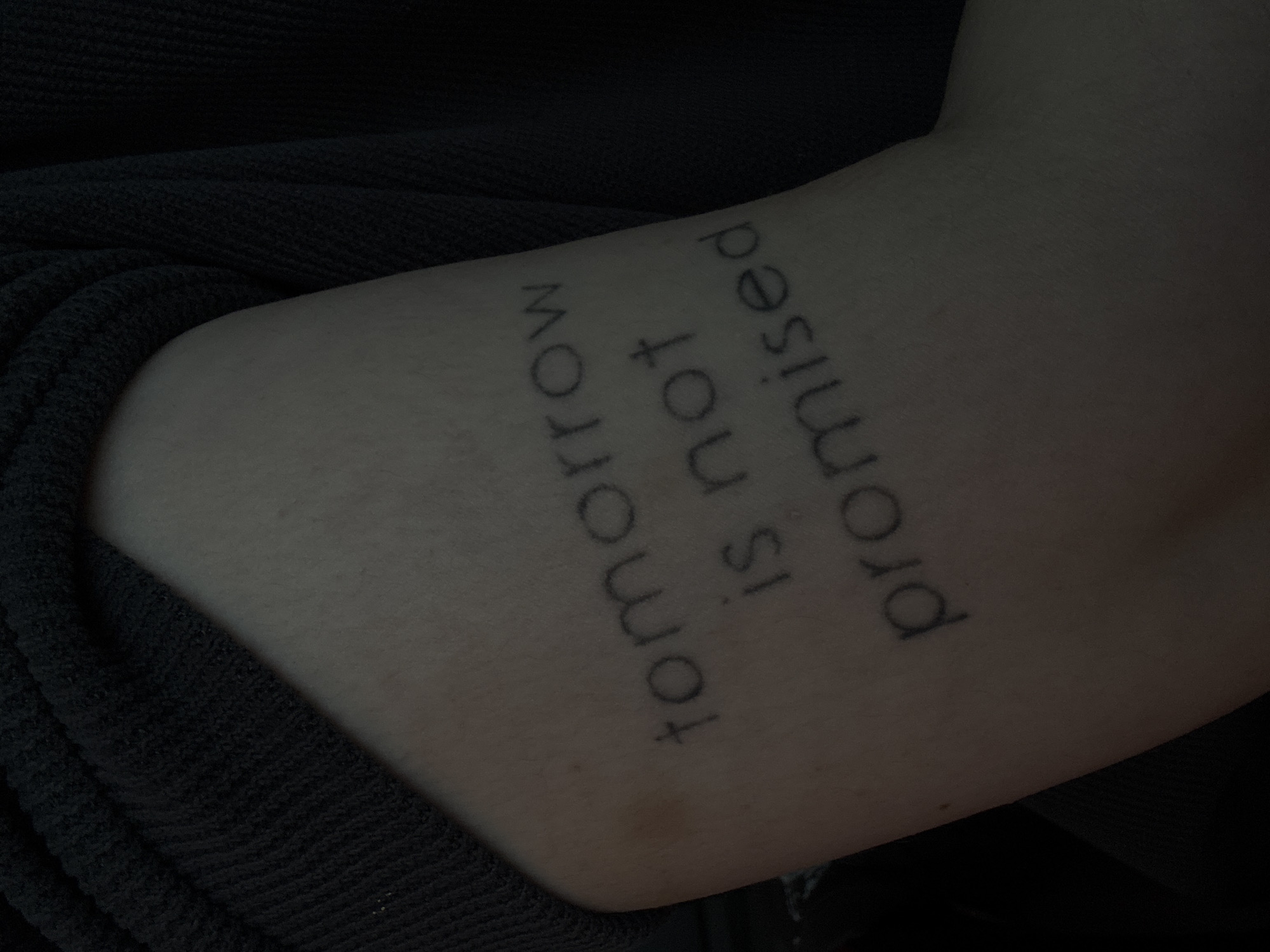 The image shows a close-up of a tattoo on a person's arm. The tattoo consists of the words "tomorrow is not promised" in a simple, sans-serif font, arranged in three lines. The tattoo is dark and appears to be relatively fresh, with some slight redness around the edges. The arm is positioned against a dark, textured fabric, possibly clothing. The lighting is dim, creating a moody atmosphere. The specific location in Saint Petersburg, Russia cannot be determined from the image.