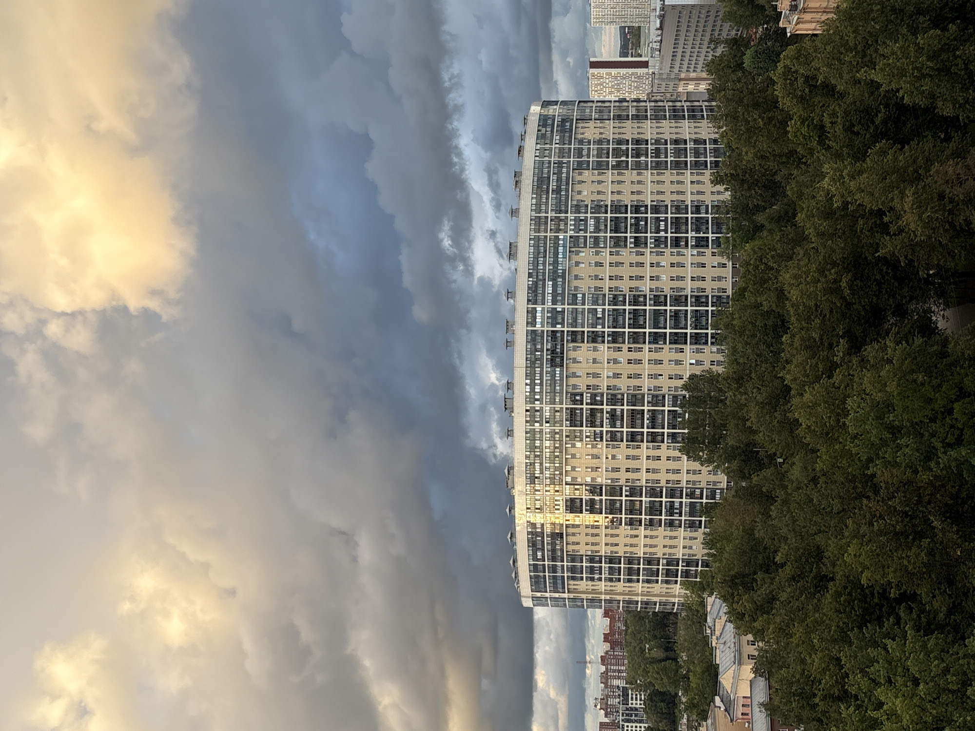 The image captures a panoramic view of the city of Otradnoye, Russia, dominated by a large, curved residential building in the foreground. The building, a modern multi-story structure, features a light-colored facade with numerous windows arranged in a grid pattern. The curved design of the building is a prominent architectural element.
Behind the main building, other structures of varying heights and architectural styles are visible, suggesting a dense urban environment. Some of these buildings appear to be residential, while others may be commercial or administrative. The distant skyline is partially obscured by the buildings and a dense canopy of lush green trees in the middle ground, which frames the lower portion of the scene.
The sky above is a dramatic display of clouds. Dark, heavy storm clouds dominate the upper half of the image, casting a brooding atmosphere. However, pockets of bright, golden sunlight pierce through the clouds, illuminating parts of the sky with a warm glow and reflecting off the windows of the main building. This contrast between light and dark clouds creates a dynamic and visually striking sky.
No people or specific actions are discernible in the image, suggesting a static, observational perspective. The scene depicts an urban landscape under a dynamic sky, likely at dusk or during a period of changing weather. The overall impression is one of a modern city coexisting with natural elements, under a sky of dramatic atmospheric conditions.