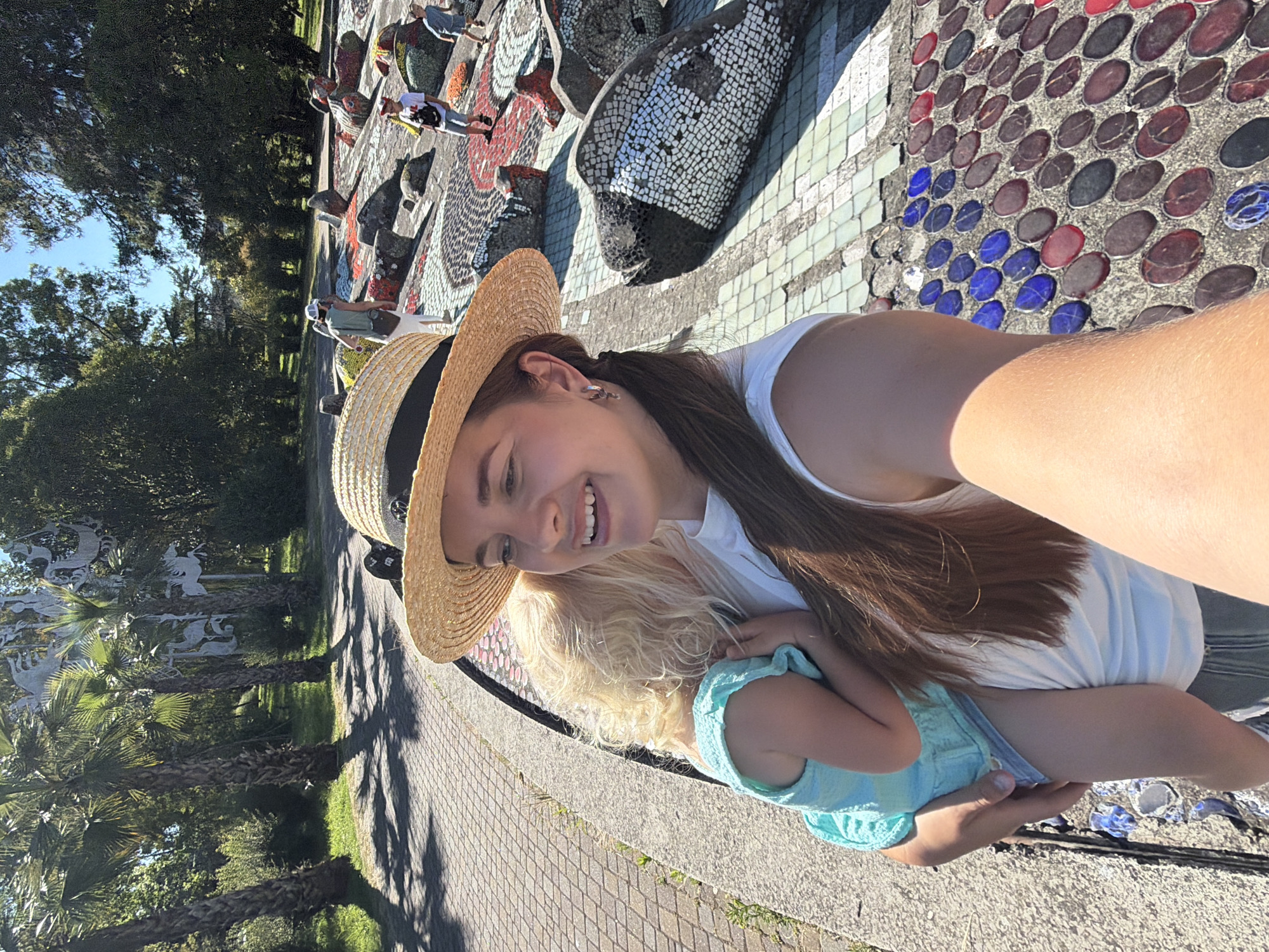 In Sochi, Russia, a woman in a straw hat is holding a young child. The woman is smiling and taking a selfie, with her arm extended. The child is wearing a light blue dress and has blonde hair. They are standing on a paved walkway next to a decorative mosaic wall. The wall is made of colorful tiles and features mosaic fish sculptures. In the background, palm trees and other foliage suggest a park-like setting. Other people, including children, are visible in the distance walking along the mosaic wall. The sun is shining brightly, casting shadows on the ground.