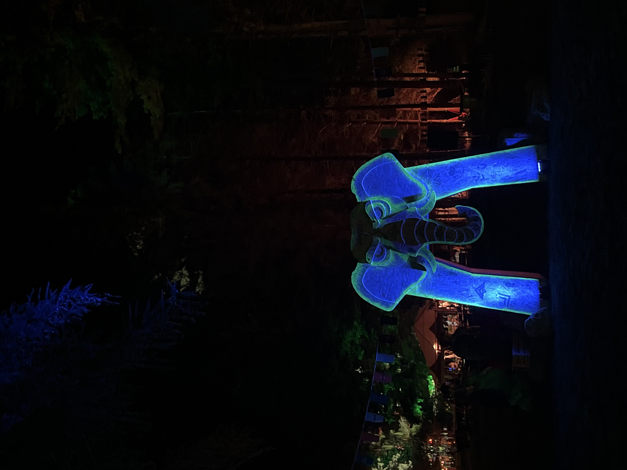 The image depicts a nighttime outdoor scene in Sremski Karlovci Municipality, Serbia, dominated by a large, illuminated elephant-shaped archway serving as a focal point. The archway, positioned centrally, emits a bright blue light with green accents outlining its features, including prominent ears, eyes, and a downward-curving trunk. Intricate patterns or glyphs are visible on the blue surfaces of the archway's support structures. An inscription resembling "DOVVELY" is visible on the elephant's forehead.
In the mid-ground to the left of the archway, an area appears to be a gathering or festival space. Several tents or stalls, illuminated with warm yellow light, are present. Multiple indistinct human figures are visible around these structures, with one figure in green attire partially discernible near the left base of the elephant archway. A string of colorful rectangular banners (blue, purple, green, red) is suspended horizontally above this area. Trees in the background to the left are illuminated with a pale green light, with some foliage in the upper left corner showing a blue hue.
To the right of the archway, a dense stand of trees forms a dark background, with some trunks and foliage partially illuminated by a reddish-orange light. A faint line of banners similar to those on the left is also present on this side. The immediate foreground consists of dark ground, with minimal detail, showing some subtle textural variations or small, dark objects near the base of the elephant structure. The overall lighting scheme suggests an artificial, decorative illumination in a natural, dark environment.