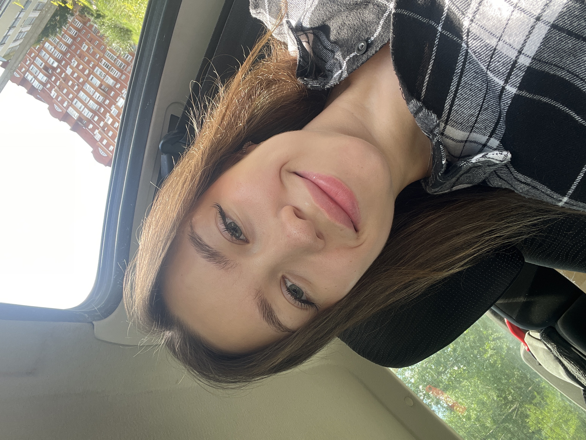 A young woman with long brown hair and green eyes is taking a selfie in a car. She is wearing a black and white plaid shirt. The background shows trees and a building, suggesting an urban or suburban environment. The image appears to have been taken during the daytime, with natural light illuminating the scene. The reflection in the car window shows a building that resembles structures found in Chaikovsky, Russia.