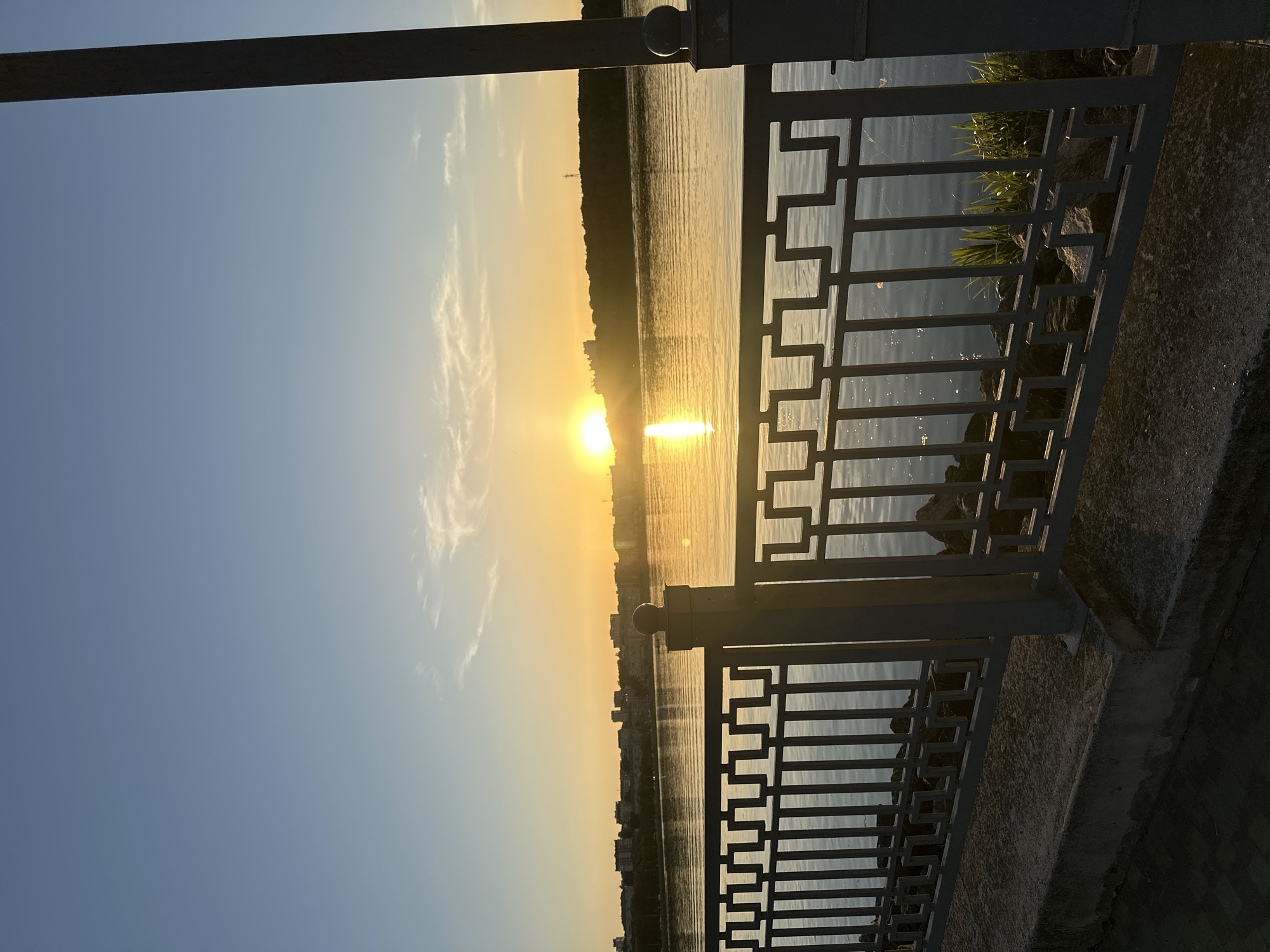 The image depicts a sunset over the Ternopil Pond, a reservoir in Ternopil, Ukraine. The sun is low on the horizon, casting a golden glow across the water and illuminating the sky with warm hues of orange and yellow. A gentle breeze ripples the surface of the pond, reflecting the light. In the foreground, a metal fence with a geometric pattern runs along the embankment, separating the path from the water. Beyond the fence, the distant cityscape of Ternopil is visible, with buildings silhouetted against the vibrant sky. The overall scene evokes a sense of tranquility and natural beauty.