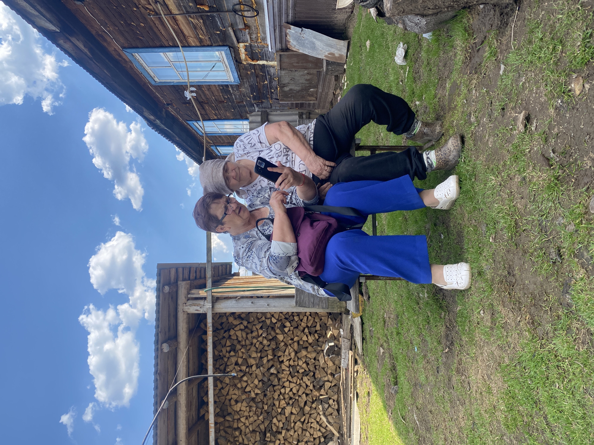 Two women are seated on a bench outdoors. One woman, wearing a white patterned shirt and a headscarf, is holding and looking at a smartphone. The other woman, wearing a light patterned blouse, glasses, and blue pants, is looking at the phone. They are sitting in front of a woodpile and an old wooden structure with a blue-framed window. The location appears to be a rural setting, with grass and muddy ground. The sky is visible, with blue sky and white clouds. Based on the information provided the location is Узяр, Russia.