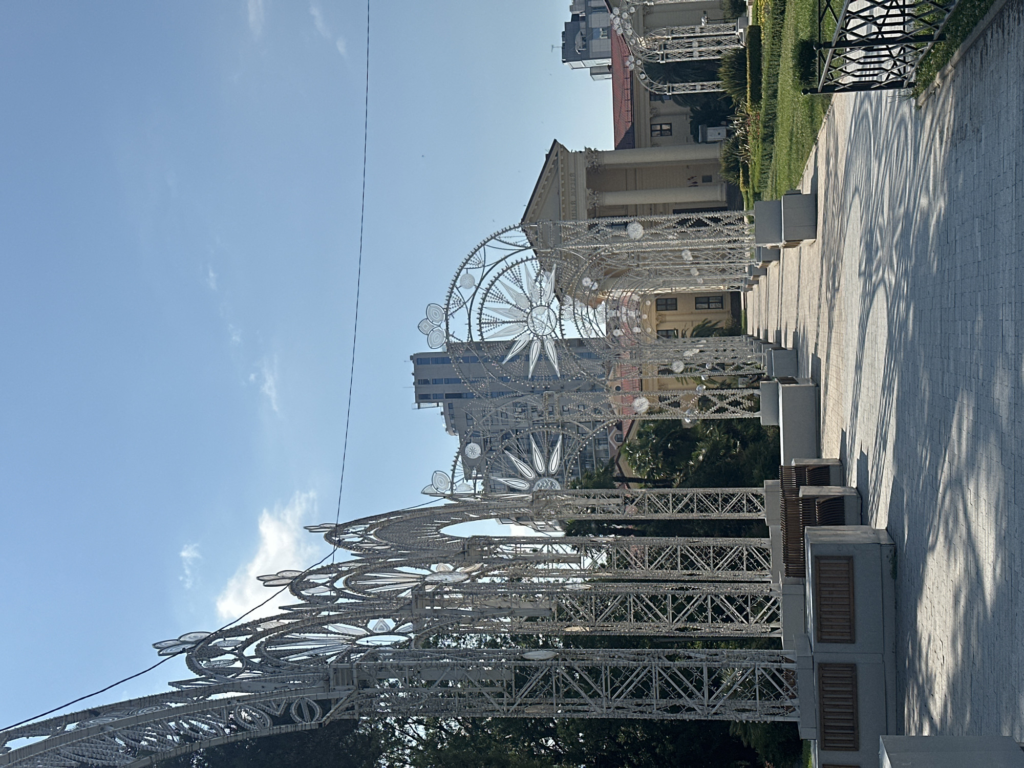 In the city of Sochi, Russia, a pathway lined with decorative white arches extends towards a grand building with classical architecture. The arches are elaborate, featuring intricate metalwork and circular decorative elements. Sunlight casts dappled shadows on the cobblestone pathway and the neatly manicured green lawn to the right. Benches are placed along the pathway, integrated into concrete bases. In the background, modern high-rise buildings rise above the classical structure. The sky is a clear blue with a few wispy clouds. The scene appears to be a public space, possibly a park or a promenade, designed for leisure and aesthetic appeal.