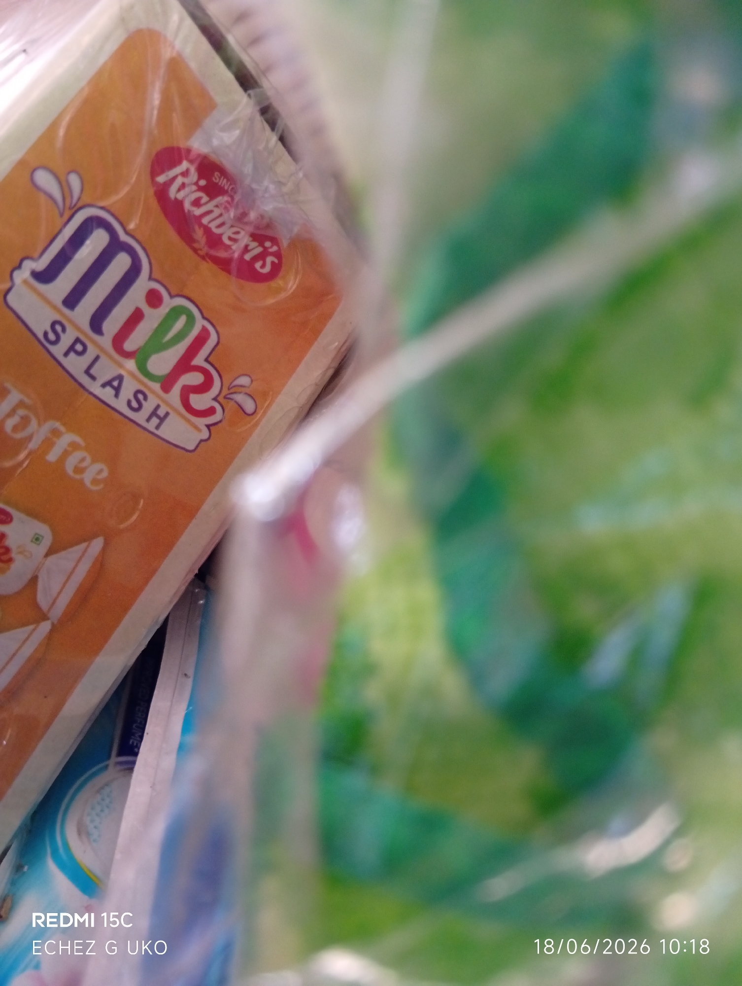 The image, captured in Jalingo, Nigeria, presents a close-up, slightly blurred view of packaged candies. The foreground features an orange wrapper with the prominent text "Milk SPLASH" in a playful font, with "Toffee" also visible. A red oval logo with "Richberri's" is present. Behind the candies, a predominantly green, out-of-focus background suggests another object or packaging. The bottom left corner displays text identifying the device as "REDMI 15C" and a location as "ECHEZ G UKO." The bottom right corner shows a date and time stamp: "18/06/2026 10:18." No people or overt actions are depicted.