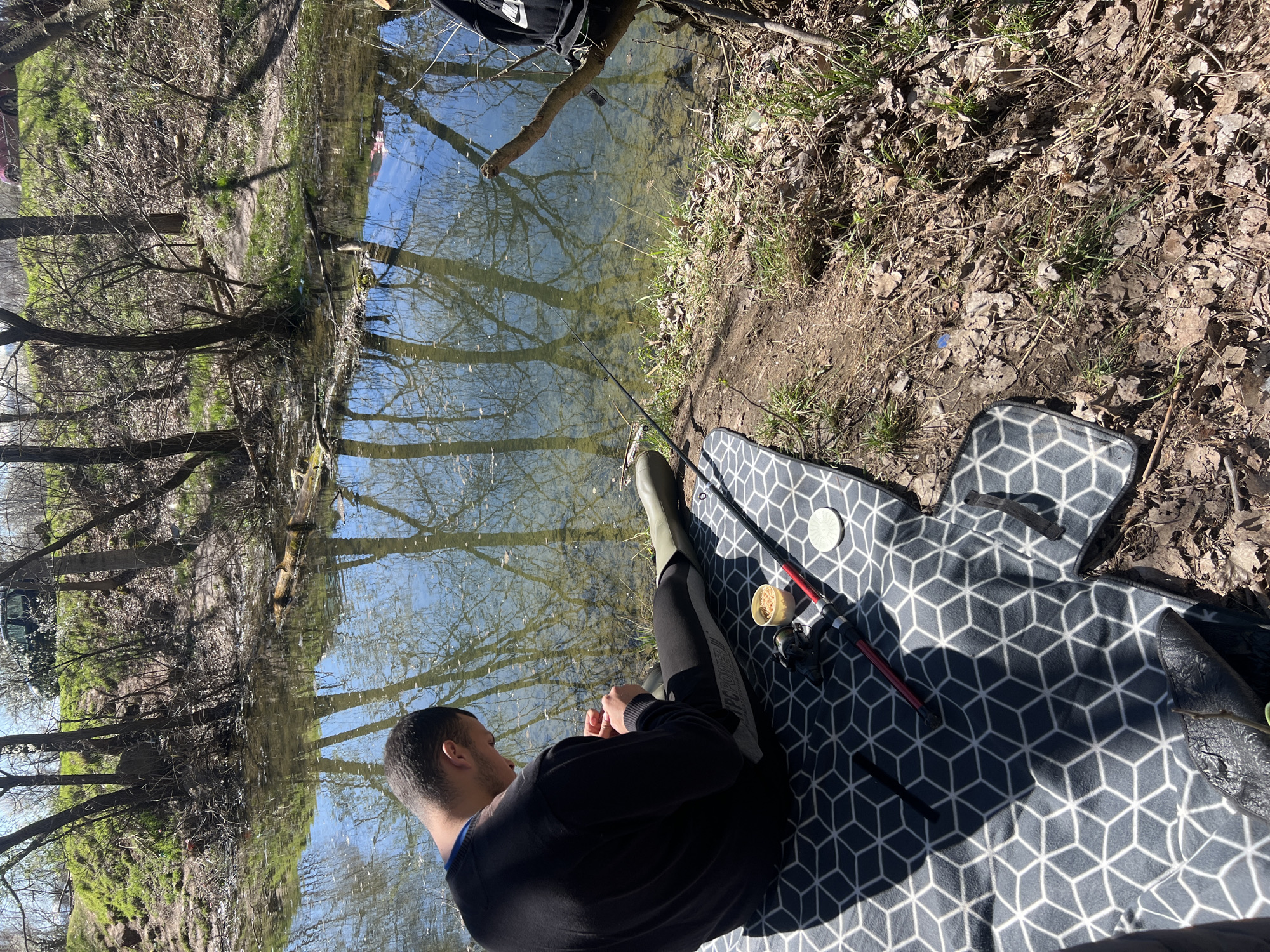 A man is fishing on the bank of a stream or river. He is wearing a black sweater, black pants with a grey stripe, and green rubber boots. He is sitting on a grey mat with a white geometric pattern. A fishing rod rests next to him, along with a small container of bait and some fishing tackle. The river reflects the surrounding trees and sky. The riverbank is covered in leaves and grass. The location is likely in Sofia, Bulgaria.