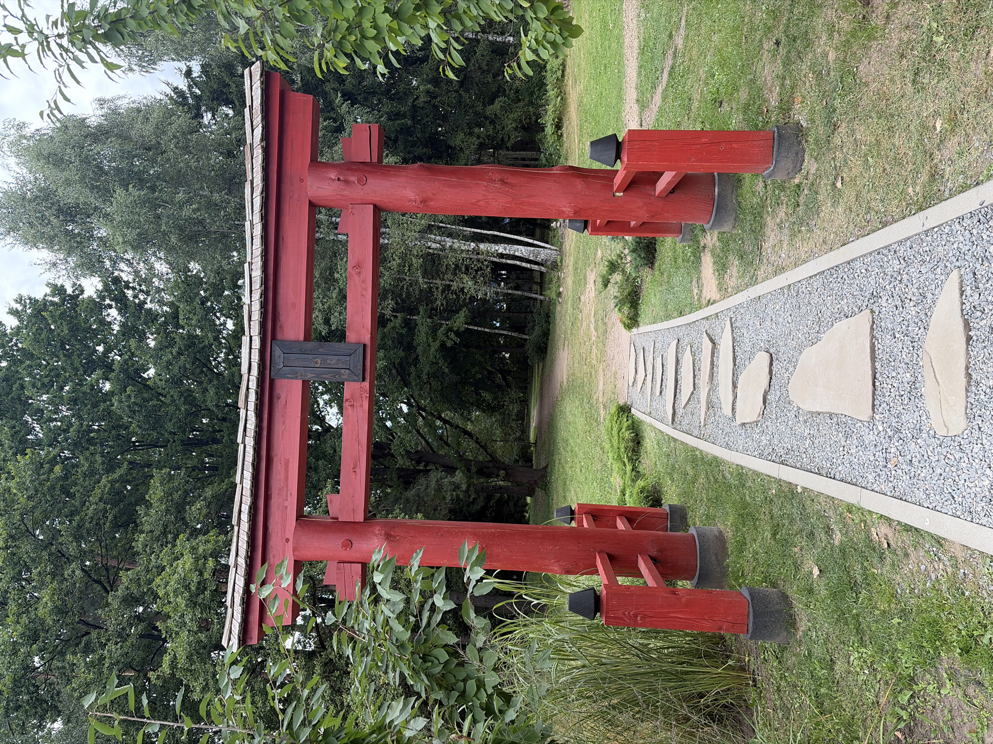 A bright red torii gate stands in a park-like setting. The gate's posts are thick and wooden, painted a vibrant red, and topped with a wooden crossbar. Above the crossbar, a multi-tiered roof is constructed with what appears to be wooden shingles. A dark rectangular plaque is affixed to the center of the crossbar. A pathway of flat, irregular stones laid on top of a bed of small gray gravel leads through the torii gate. The pathway is bordered by a thin strip of concrete on the right. To the left of the pathway and gate are tall, thin green grasses and lush green foliage from trees. To the right of the gate, the pathway continues into a grassy area with more trees in the background. The ground is covered in green grass, with patches of dirt and fallen leaves visible. The background is filled with a dense forest of various trees, their leaves a deep green. The sky is overcast with diffused light. There are no people or animals visible in the image. The scene appears to be a serene and natural setting, possibly a garden or park designed with Japanese influences.