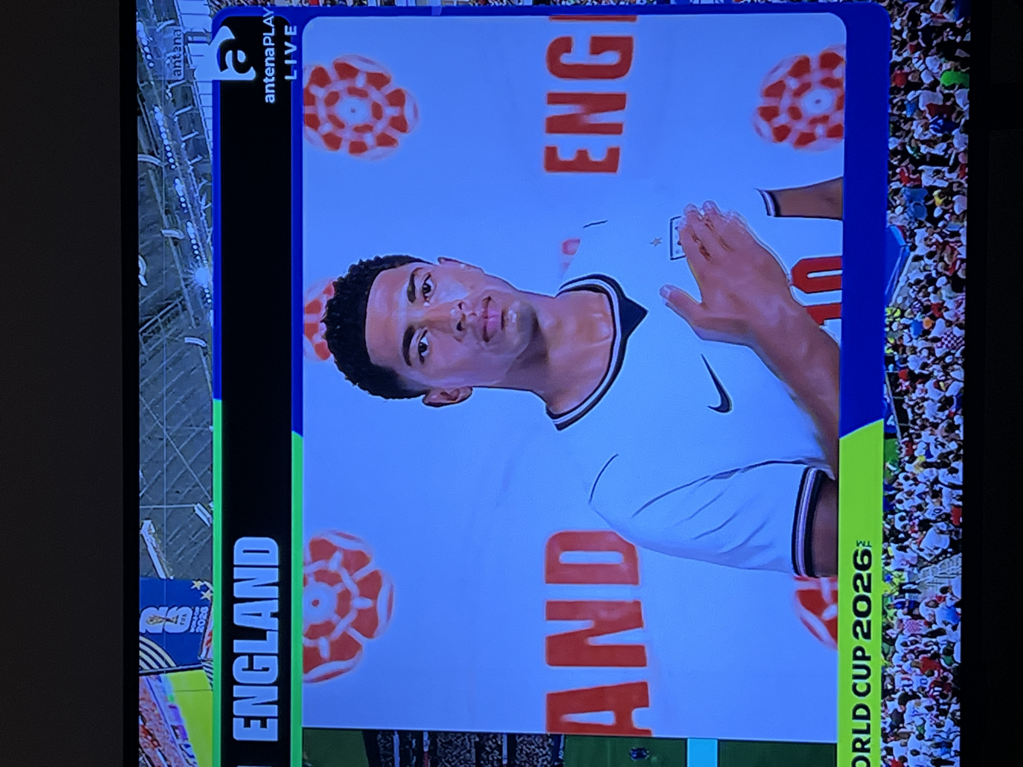 The image shows a television screen displaying a promotional image for the "WORLD CUP 2026". A male athlete, wearing a white jersey with a Nike logo and the number 10, is prominently featured. He is placing his hand over his chest. The background is light blue with several red and white patterned circular motifs, resembling roses or stylized footballs. The words "ENGLAND" are displayed in large blue letters on a black bar at the top of the screen, and "AND" and "ENG" are visible in large red letters on the blue background. In the top right corner, there is a logo for "antenaPLAY LIVE". Below the athlete, a crowd of spectators is visible. The image is framed by a television bezel. The location in Florești, Romania is not discernible from the provided image.