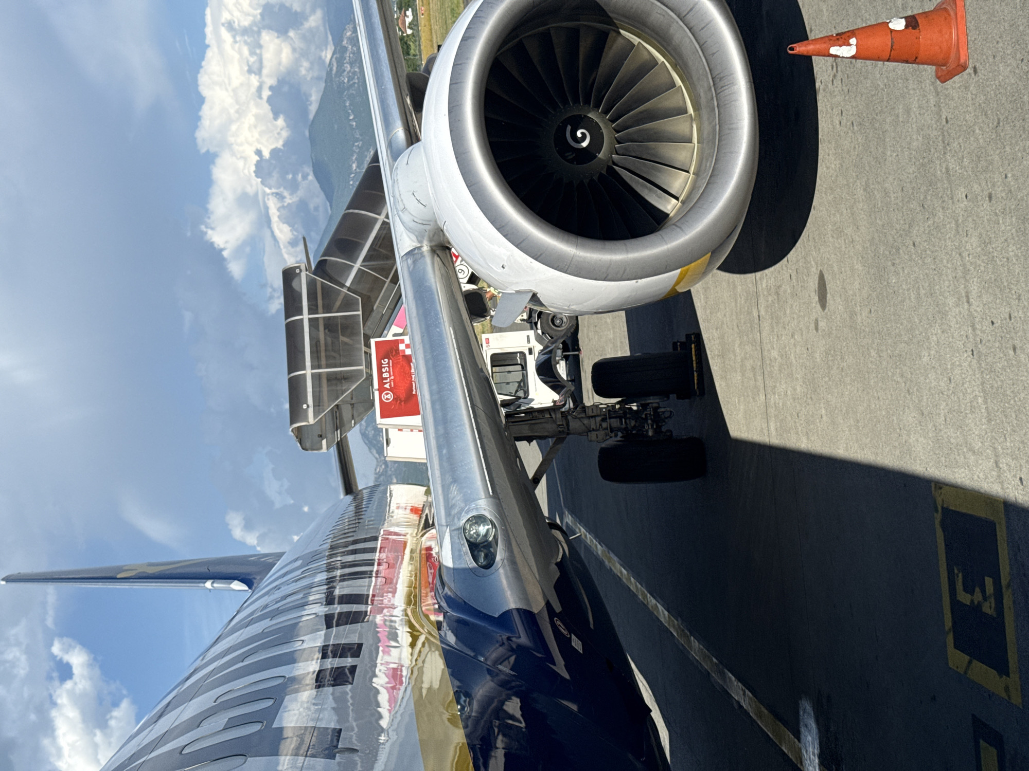 The image shows a close-up view of the engine and wing of an airplane on the tarmac at an airport. The airplane is a large commercial jet with a distinctive blue and silver livery. The engine is a turbofan, with visible fan blades inside the cowling. A jet bridge is connected to the aircraft, and a ground vehicle, possibly a baggage tug, is parked nearby. In the background, mountains are visible under a cloudy sky. The scene appears to be set during the daytime, with strong sunlight casting shadows. The ground is asphalt, marked with yellow lines and the letter "E" painted in a dark blue rectangle. An orange traffic cone is visible in the foreground. The location is identified as Kruja, Albania.