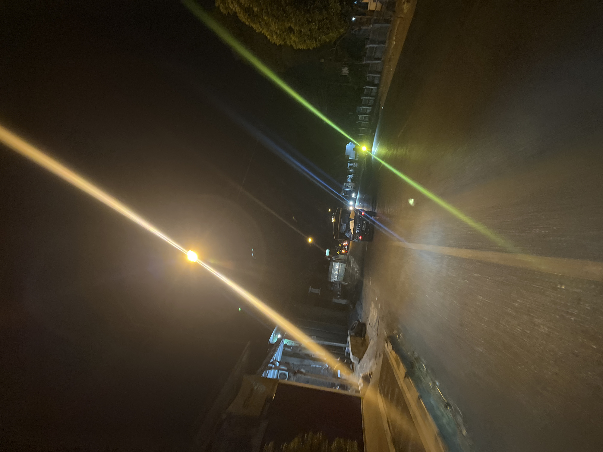 The image depicts a nighttime scene on a street, possibly in Lebuawu, Indonesia. A vehicle, appearing to be a utility truck or a small bus, is driving away from the camera. The street is wet, reflecting the light from street lamps and the vehicle's headlights, suggesting recent rain or a damp surface. Along the left side of the road, there are buildings with closed storefronts and some debris, including what appears to be a discarded tire. On the right side, a tree line and a fence are visible. Bright streaks of light, characteristic of lens flare from streetlights, cut across the image from various angles, with distinct yellow and green hues. The overall lighting is dim, typical of a street at night, with localized illumination from the street lamps.