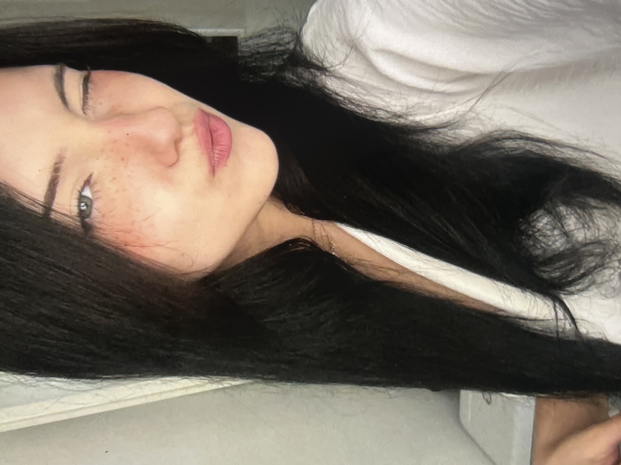 The media file is a portrait of a young person with long, dark hair. The person is lying down, with their face turned towards the camera. They have fair skin with freckles, light green eyes, and pink lips. Their eyebrows are well-defined, and they are wearing a white garment that appears to be a shirt or jacket. The background is blurred and consists of a textured white surface, possibly fabric or bedding. The lighting suggests an indoor setting, and the time of day is not clearly indicated. The overall mood of the image is relaxed and casual. There is no visible text in the media file.