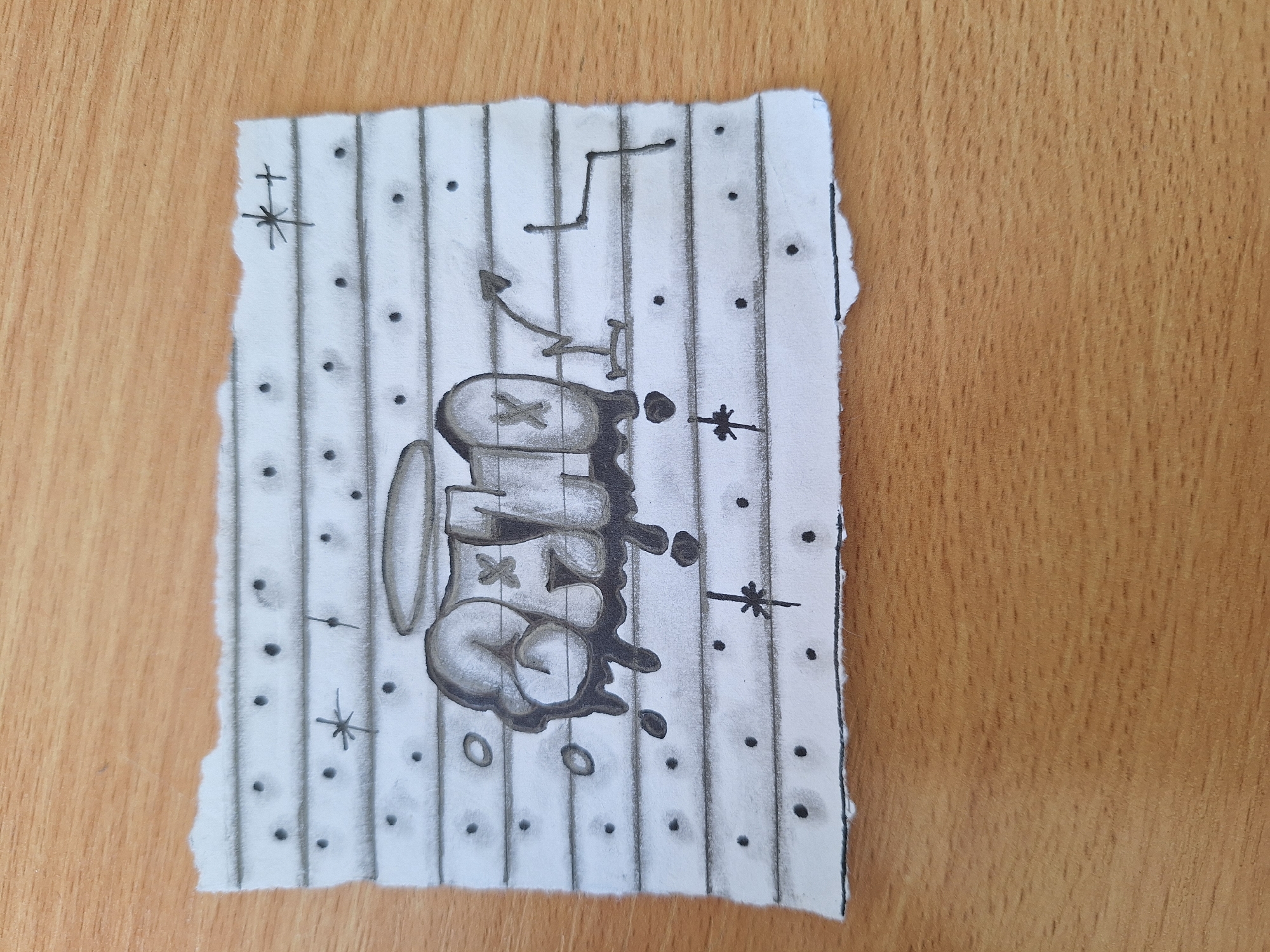 A torn piece of lined paper with a graphite drawing of graffiti is placed on a wooden surface. The graffiti appears to be a stylized word or name with shading to give it a 3D effect, and dripping elements. Small dots and star-like shapes are also drawn on the paper, resembling a starry night or decorative elements. There are no people or specific Varna landmarks visible in the image. The drawing itself is the sole subject.