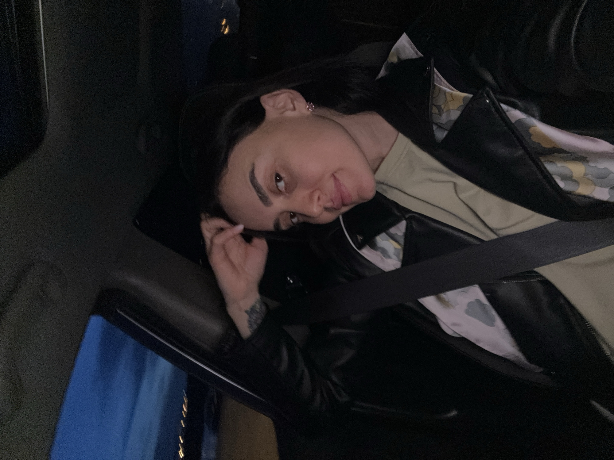 A young woman is captured in a casual, close-up shot from inside a vehicle at night. She lies with her head tilted towards the camera, offering a slight smile or smirk. Her dark hair is swept back, revealing a small, pinkish earring in her right ear. She is dressed in a black leather jacket, worn over a light green or khaki t-shirt. The jacket's inner lining features a distinctive pattern of light blue and grey clouds against a pale yellow background. A dark seatbelt diagonally crosses her chest, indicating she is seated and secured. Her right hand, adorned with a tattoo on the wrist (appearing to be a floral or abstract design) and light-colored nails, is positioned near her head.
The setting is dimly lit, with the interior of the vehicle appearing dark. Through the window, a blurry blue light is visible, suggesting urban illumination or artificial light sources outside. Faint, warmer yellow lights can also be seen in the distant background. The overall impression is relaxed and candid, likely taken during a nocturnal journey in Tbilisi, Georgia. No visible text is present in the image.