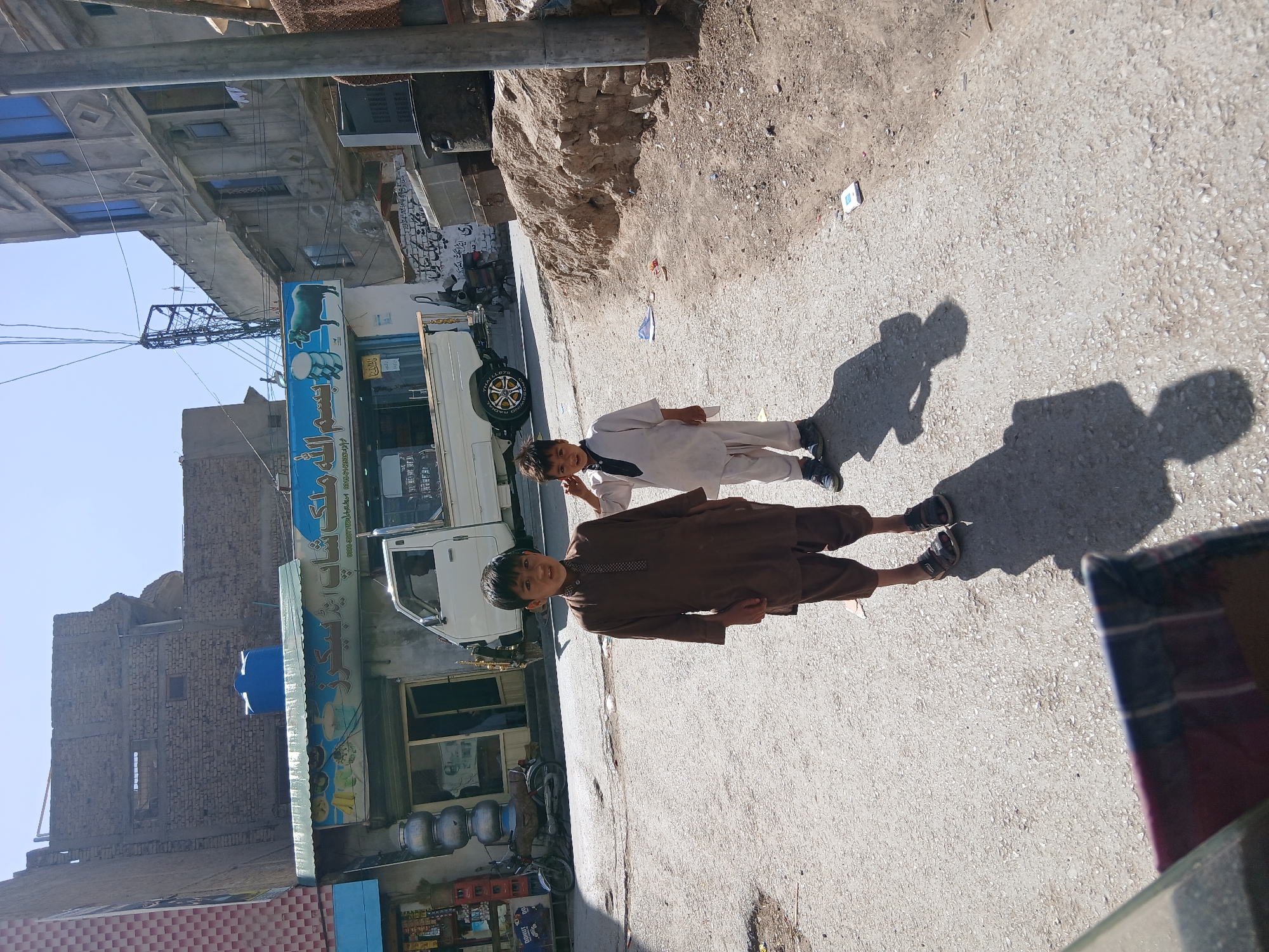 This image, taken in Quetta City Tehsil, Pakistan, captures two young boys standing on a sunlit, paved street. The scene is oriented with the clear, bright blue sky at the top and the street at the bottom, illuminated by strong sunlight that casts long shadows.
The boy closer to the camera, appearing to be around 8-10 years old, wears a dark brown shalwar kameez and black shoes. He looks directly at the viewer with a faint smile. Behind him stands another boy, slightly older (perhaps 10-12 years old), dressed in a white shalwar kameez and black shoes, also facing the camera with his hands clasped. Both boys cast long, distinct shadows on the gravelly asphalt road, indicating either early morning or late afternoon sun. Another shadow, likely belonging to the photographer, is visible in the bottom right foreground.
On the left side of the street, a bustling urban environment unfolds. A white pickup truck, possibly a light commercial vehicle, is parked near a shop. Its tire distinctly shows the text "DUNLOP RADIAL." The shop features a prominent blue signboard adorned with an illustration of a bull and a barrel, and visible Urdu text. The main readable part appears to be "ہاشم دال چنا" (Haashim Daal Chana), suggesting a food or eatery business. Below this, stacks of blue plastic barrels or containers and red crates, one visibly marked "BUBBLY," are arranged outside the shop.
Beyond the shop, a multi-story concrete building with blue-tinted windows and balconies rises. Further in the background, another brick building with a blue water tank on its roof is visible. Electrical wires crisscross above the street, and a metal utility structure stands against the sky. The ground consists of a mix of paved road and rougher, dusty terrain near the buildings, with some scattered litter, including small white paper. The overall impression is a lively street scene in a South Asian city during a clear, sunny day. The boys appear calm and seem to be posing for the camera.