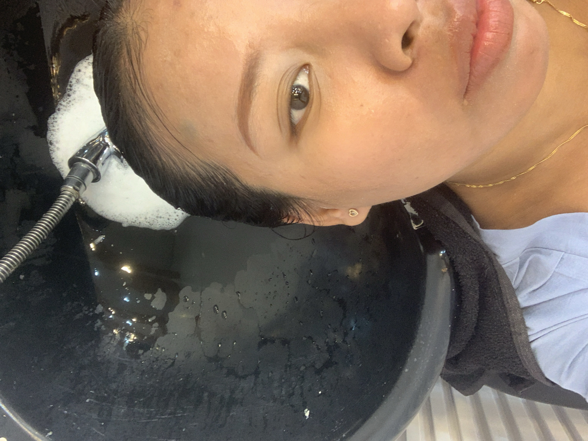 The image shows a close-up of a person's face, focusing on the right side of their face and eye. The person is positioned next to a large, dark sink, which appears to be a wash basin, possibly in a salon or spa. A silver showerhead with a flexible hose is visible above the sink, with white foam emanating from it. The person is wearing a light blue shirt and a delicate gold chain necklace. Their right ear is adorned with a small, heart-shaped earring. The background shows the interior of a facility with what looks like tiled walls. The overall scene suggests the person is undergoing a hair washing or treatment process in Denpasar, Indonesia.