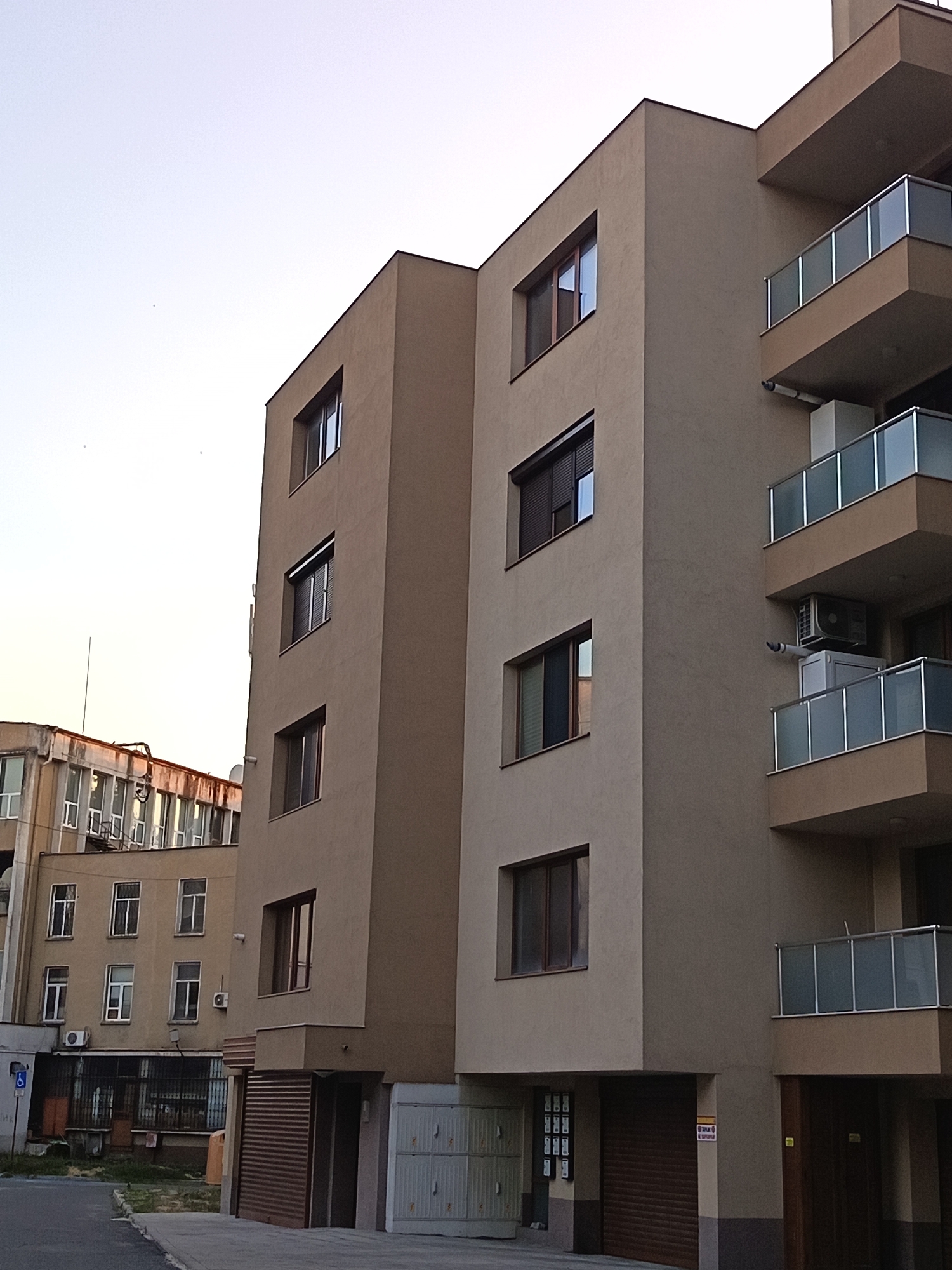 This is a photograph of a modern apartment building with a tan facade and glass-paneled balconies, located in Yambol, Bulgaria. Adjacent to it, a portion of an older, lighter-colored building with multiple windows and visible pipework can be seen. The sky is a pale, overcast blue. In the foreground, a paved area leads to the entrance of the modern building, which features a brown roll-up door and a white, multi-panel garage door. A blue and white sign with a wheelchair symbol is visible on the left side of the image. The scene appears to be a residential area.