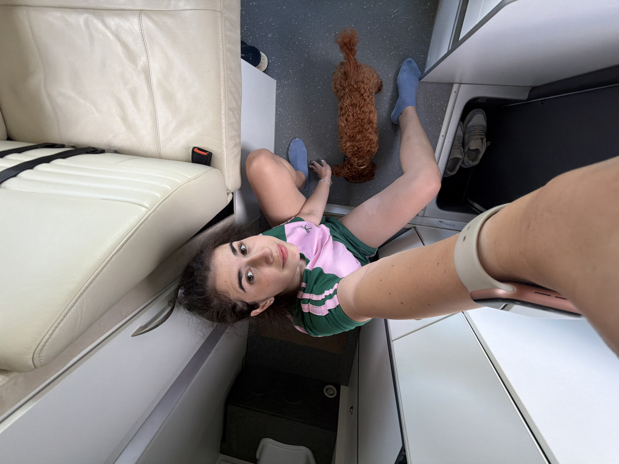 The image depicts a woman and a small dog in what appears to be the interior of a vehicle or private aircraft. The woman is seated on the floor, her legs bent and extended. She is wearing a green and pink t-shirt and blue socks. A small, reddish-brown dog, possibly a poodle mix, is also on the floor, near the woman's legs. In the background, there are cream-colored leather seats with black seatbelts, suggesting a luxurious interior. The floor is a dark, speckled material. There are also some storage compartments visible. The scene is captured from a high-angle perspective, as if the photographer is holding the camera above their head. The location in Nea Moudania, Greece is not discernible from the image itself.