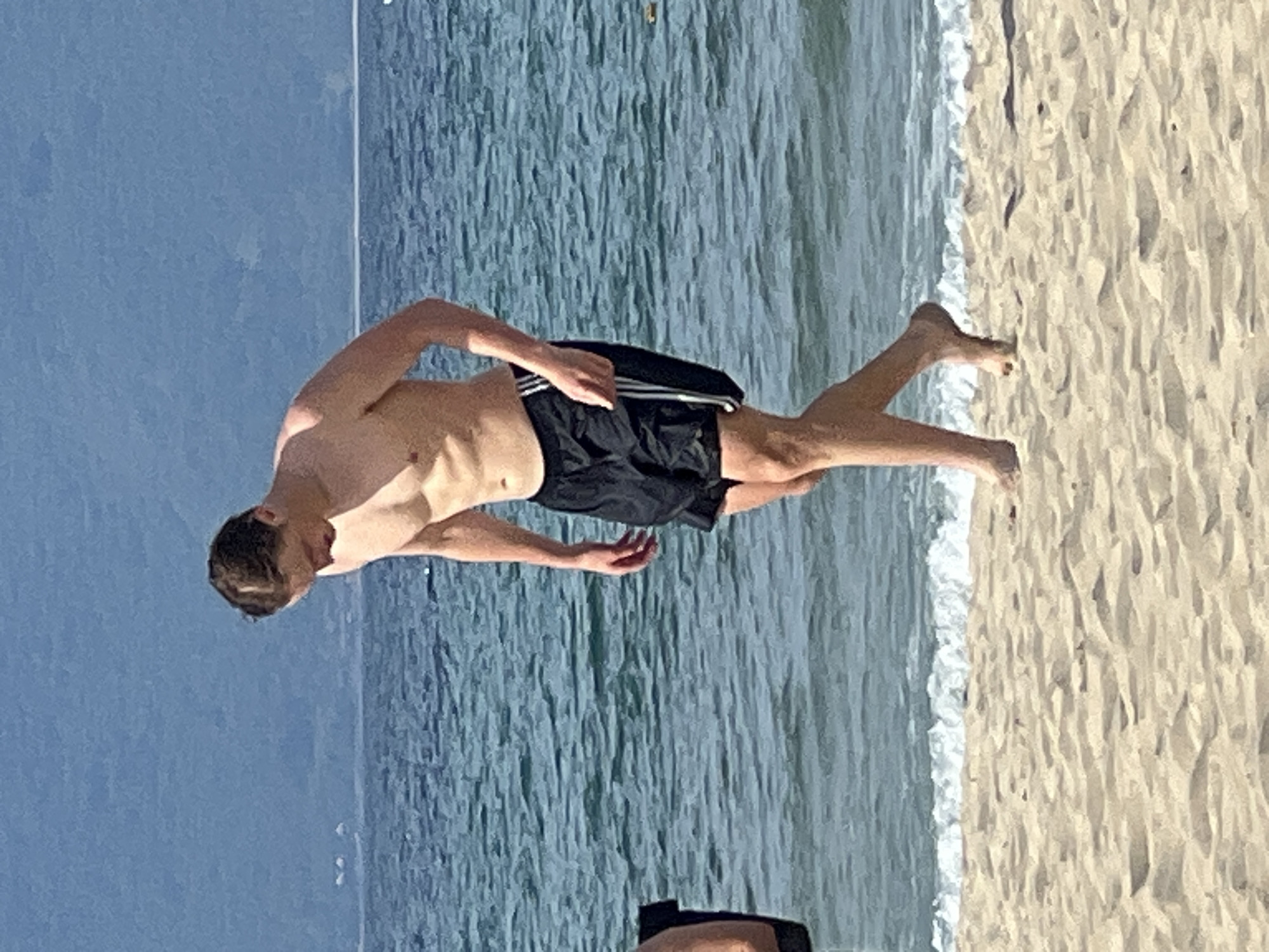 A young man with short brown hair, shirtless and wearing black swim trunks with white stripes down the sides, walks along a sandy beach. He is looking down towards the sand. The ocean, with gentle waves and a blue hue, is behind him, and in the distance, a hazy coastline is visible under a pale blue sky. The scene appears to be a typical sunny day at a beach in Đà Nẵng, Vietnam.