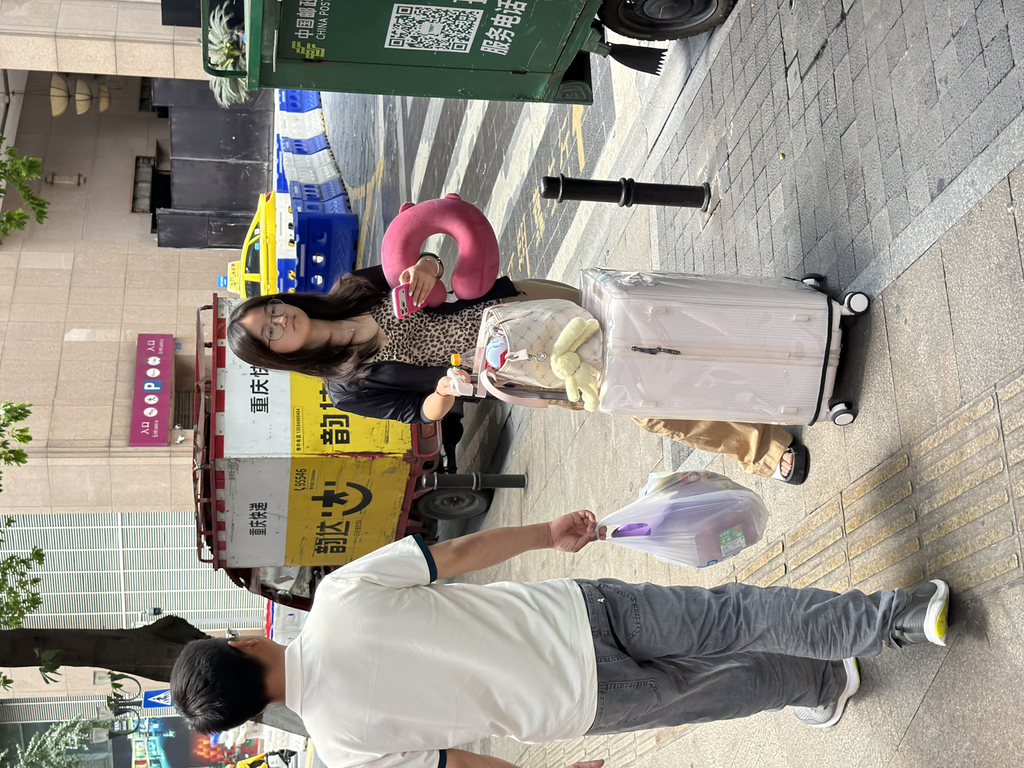 In Yuzhong District, China, on a paved sidewalk, a man wearing a white t-shirt and grey jeans walks away from the camera, carrying a transparent plastic bag filled with items. To his right, a young woman wearing glasses, a leopard-print top, and a black jacket stands next to a light pink suitcase. She is holding a pink neck pillow in her left hand and a smartphone in her right. A beige shoulder bag with stuffed animal keychains rests on top of the suitcase. In the background, a yellow taxi is visible, along with a yellow and white truck labeled "重庆快递" (Chongqing Express). A green China Post vehicle is parked on the right side of the frame. The environment suggests an urban street scene with pedestrian and vehicular traffic.