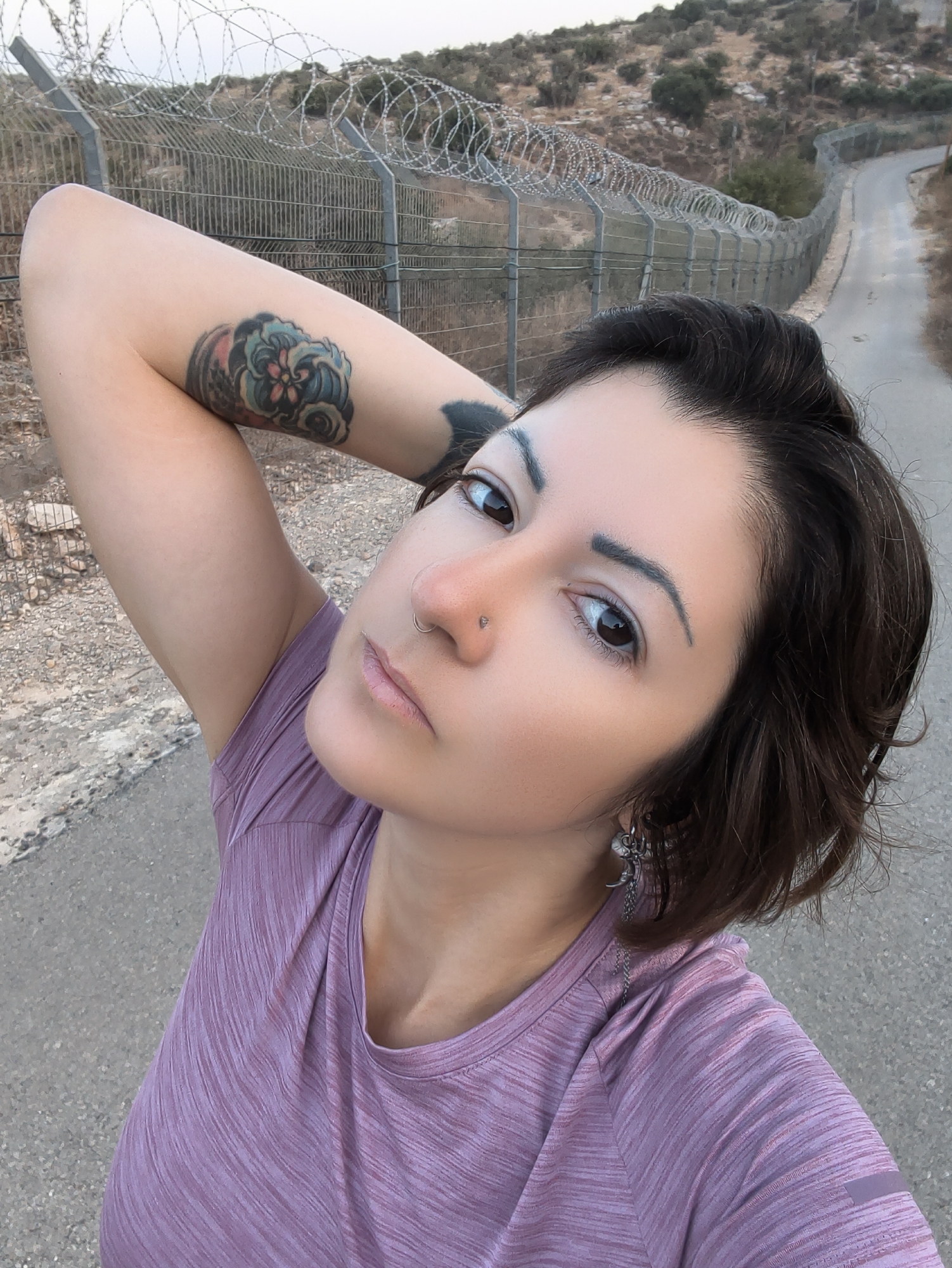 A woman with dark hair and tattoos on her arm is taking a selfie outdoors. She is wearing a purple t-shirt and an earring. She has a nose ring and her eyebrow is arched. The background shows a barbed wire fence and a road leading up a hill. The location is the Lachish Regional Council, Israel.