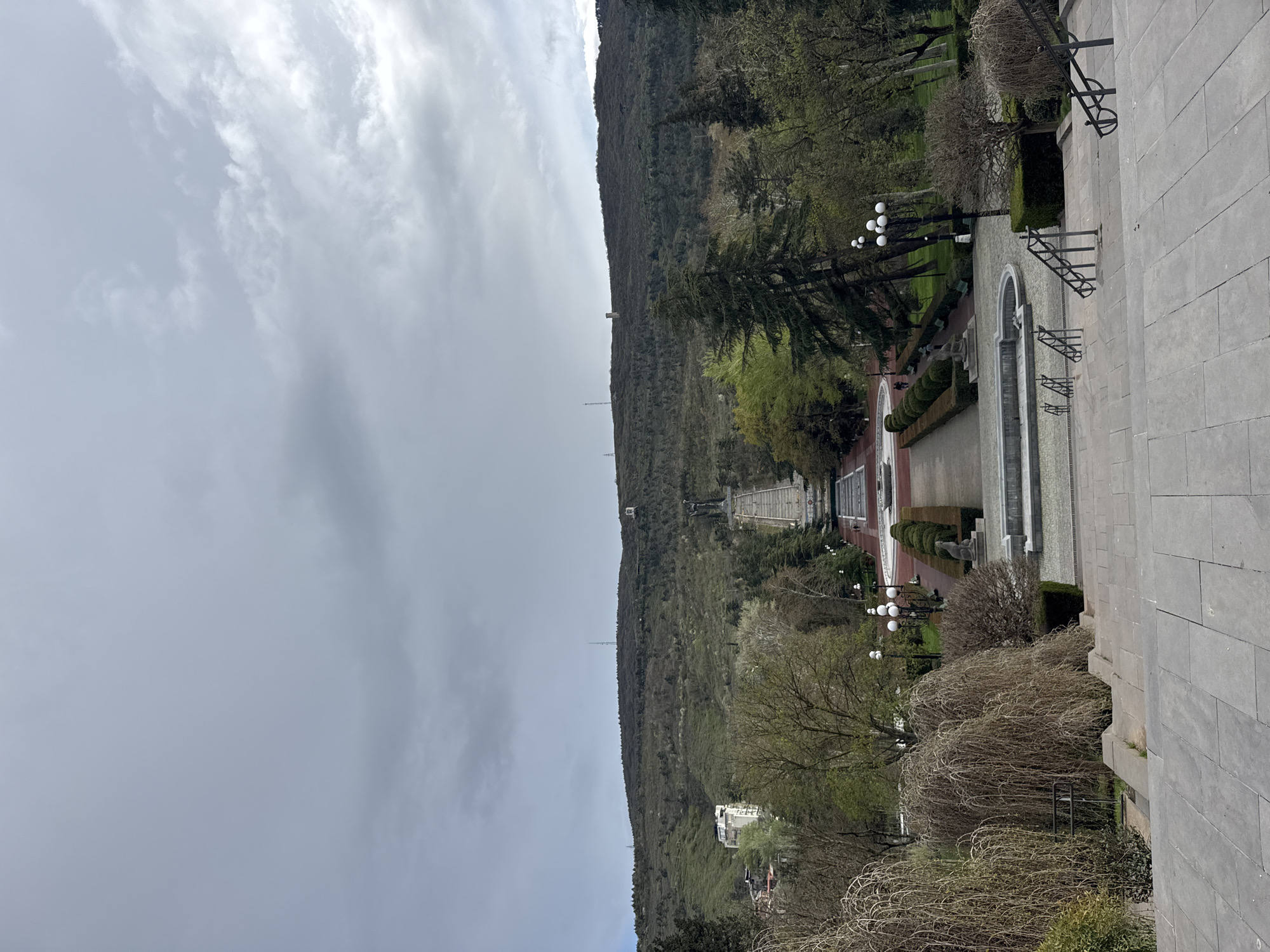 The image shows a cityscape in Tbilisi, Georgia, focusing on a long, symmetrical pathway leading up a hillside. The pathway is flanked by manicured hedges, trees, and evenly spaced lampposts. At the top of the path, atop the hillside, there appears to be a monument or statue. The foreground shows a section of gray stone paving, suggesting a high vantage point from which the photo was taken. The sky is overcast with gray clouds. The hillside is densely vegetated, and a few buildings are visible among the trees at the base of the hill.