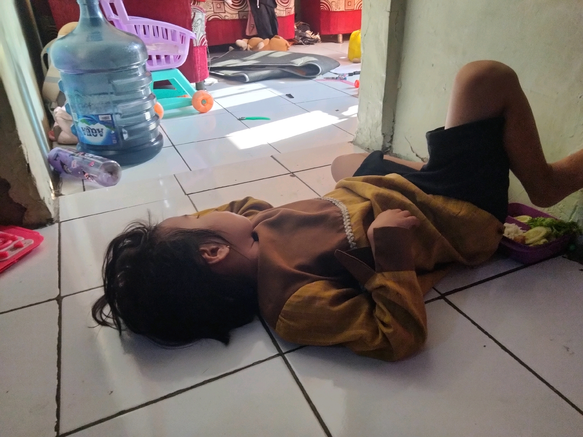 The image shows a young child lying on their side on a tiled floor in what appears to be an indoor living space. The child is wearing a yellow and brown patterned garment and black shorts. Their head is resting on the floor with their hair spread out. In the background, to the left, is a large blue water cooler bottle. Further back, there is a child's toy shopping cart with a purple basket. The floor is tiled in white with dark grout lines. Sunlight is casting shadows across the floor from the right side of the image. A person's leg, wearing black shorts, is visible on the right side of the frame, suggesting the photographer or another individual is present. On the far right, a purple container holding food, possibly salad or fruit, is visible. The environment appears to be a home in Suka Sari, Indonesia, given the context of the location.