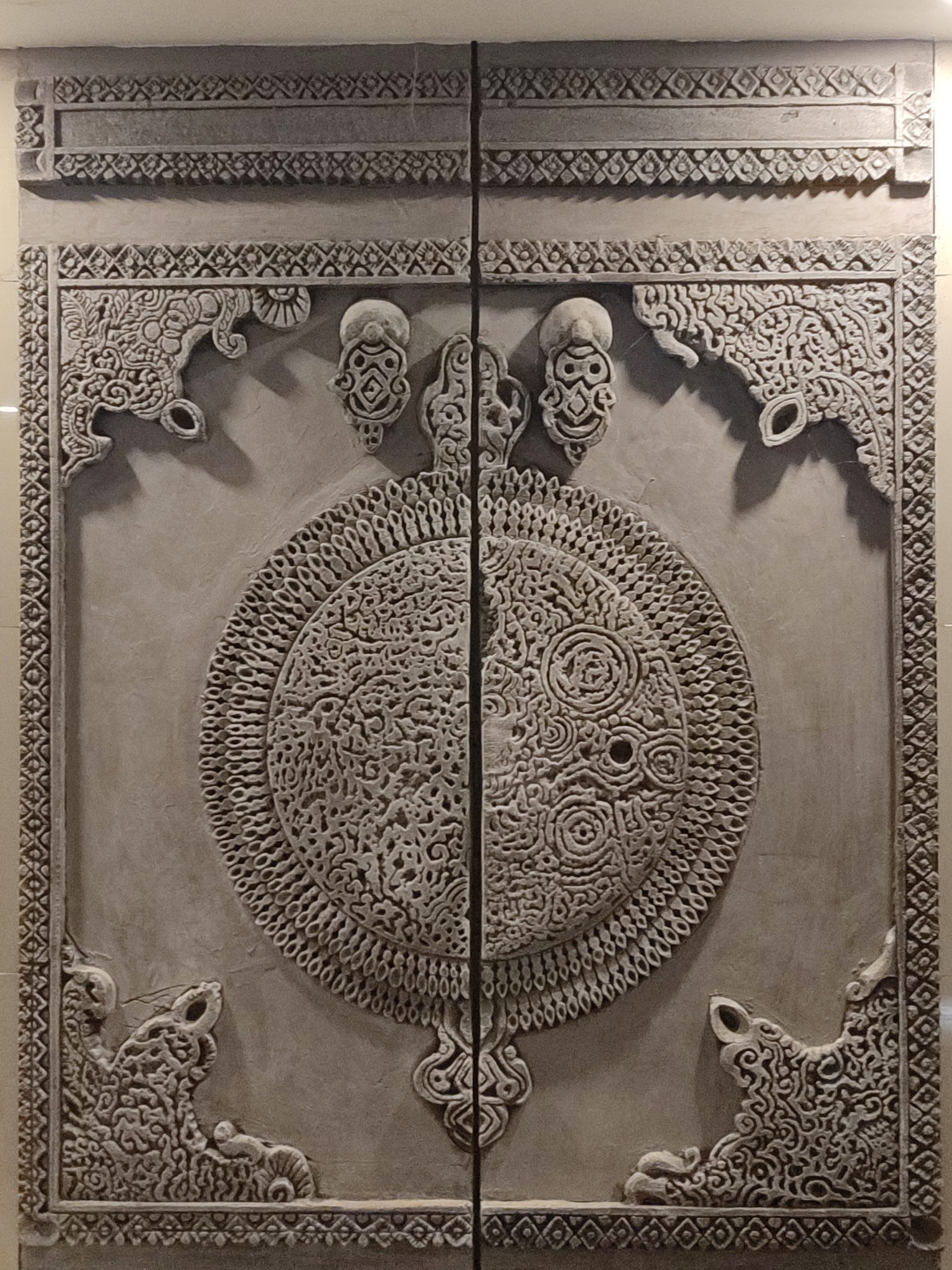 The image depicts two intricately carved gray panels, possibly doors or decorative wall pieces. The panels are adorned with a central circular motif featuring complex, swirling patterns. The upper portion of each panel includes symmetrical, ornate designs, including what appear to be stylized animal or mythical creature representations in the corners and smaller anthropomorphic figures near the center. The edges of the panels are bordered with a repetitive, geometric pattern. While the precise location is difficult to determine without additional context, the intricate carvings suggest a cultural or religious significance. The city of Muang Pattaya, Thailand, is referenced in the request but not depicted.