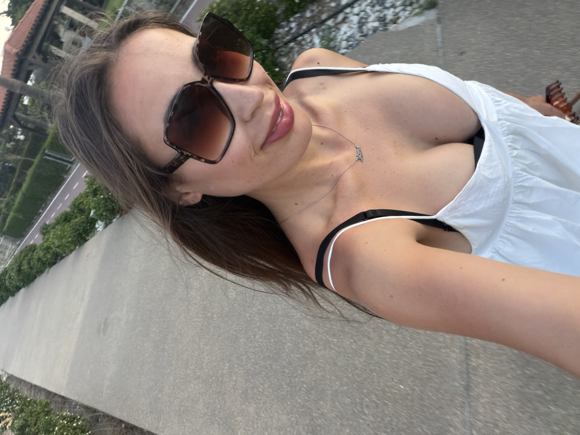 A high-angle selfie captured outdoors in Meneou, Cyprus, depicts a woman from the chest up, positioned slightly off-center to the right, facing towards the viewer with a slight downward gaze. She has long brown hair, visibly windblown. Her face is partially obscured by large, square, tortoiseshell-patterned sunglasses with gradient lenses. She is smiling, revealing pink-tinted lips. She wears a white, deep v-neck sundress or top with wide straps, beneath which black bra or bikini top straps are visible. A silver necklace with a "Mama" pendant hangs around her neck. Her right arm, visible on the lower right, holds a brown hair clip or comb. The background features a light gray concrete pathway, bordered by green foliage. Further back, a narrow paved road or pathway with white dashed lines is visible, alongside more greenery and a building with a red-tiled roof and columns, possibly a pavilion or pergola. The lighting suggests late afternoon or early evening.