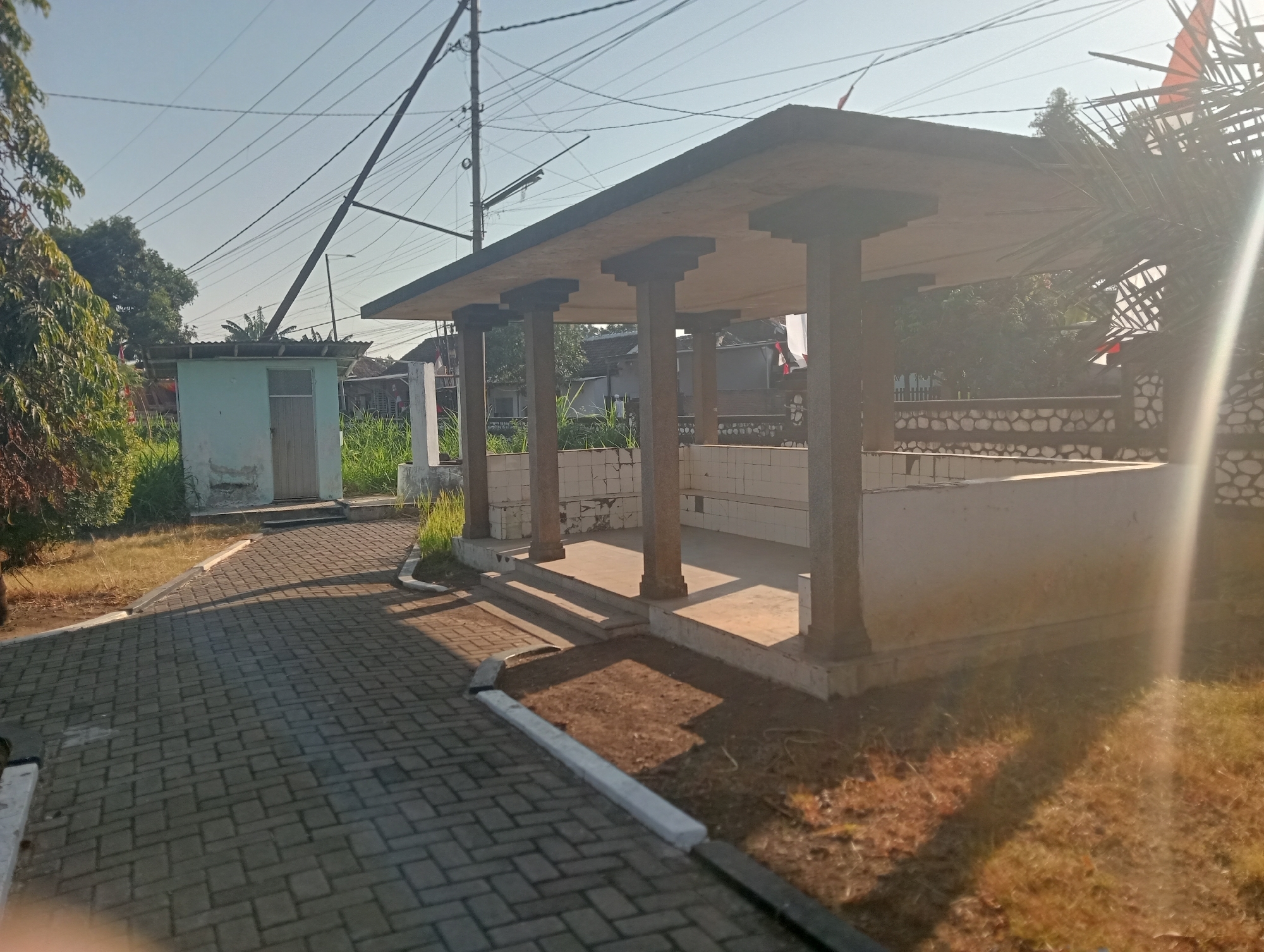 The image shows an outdoor scene on a sunny day, likely in the city of Bloro, Indonesia. In the foreground, a paved walkway made of gray interlocking bricks curves to the left. The walkway is bordered by grass and a low concrete curb.
Dominating the midground is a covered structure with a flat, dark roof supported by four stone pillars. This structure appears to be an open-air pavilion or seating area. Inside, there are white tiled benches along the back wall, and a white tiled floor. Steps lead up to the pavilion.
To the left of the pavilion, a small, light blue building with a grey metal roof and a wooden door is visible, surrounded by tall green grass.
In the background, there are houses with red tile roofs, more greenery, and utility poles with numerous electrical wires crisscrossing the sky. The sky is clear and blue. To the right, a palm frond is visible, catching the sunlight, and there is a glimpse of a red and white flag on a pole. The overall impression is one of a simple, somewhat weathered public space.