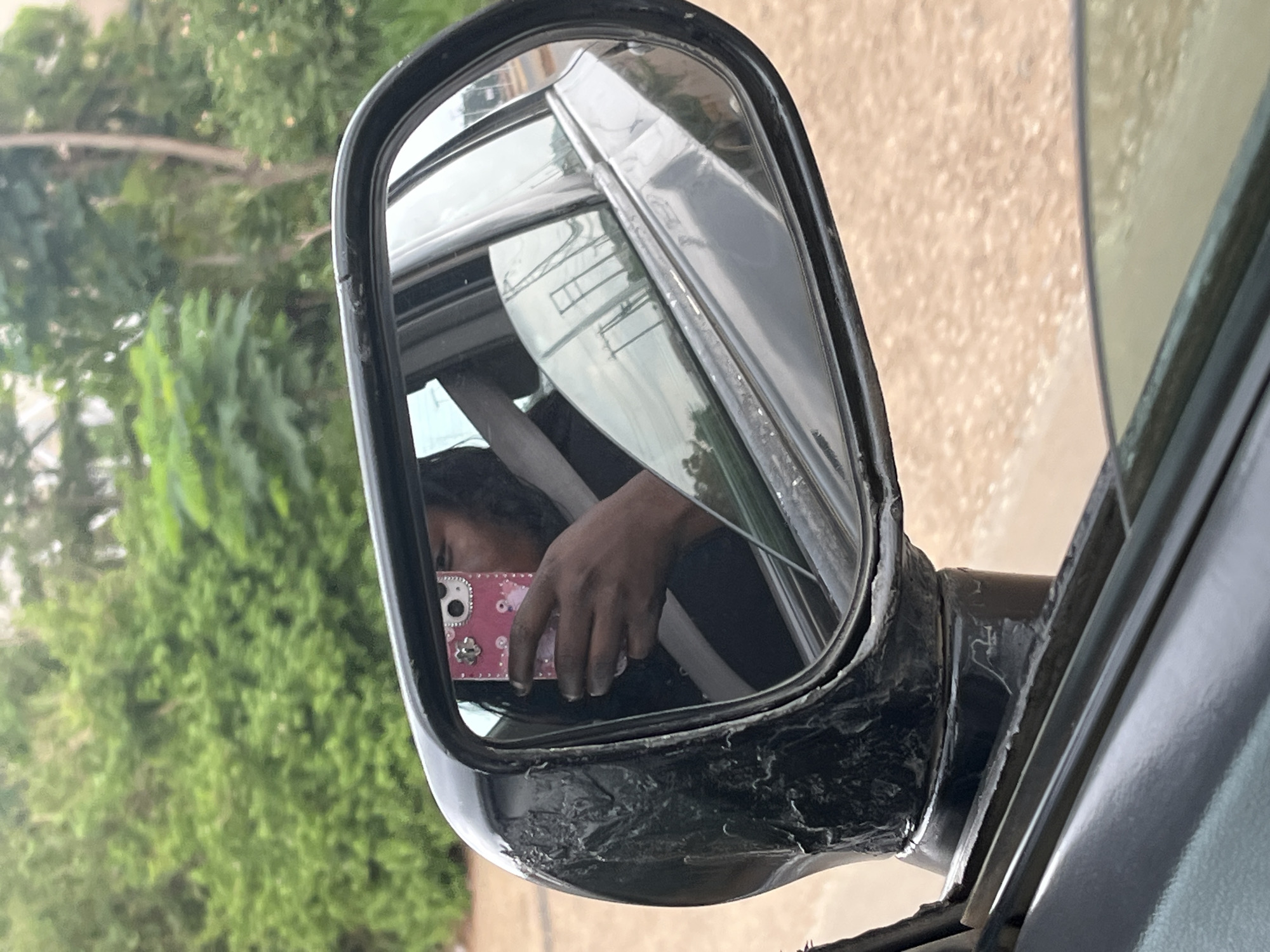 The image depicts a car's side mirror, reflecting a person taking a picture with a pink phone. Outside the car, green foliage is visible. The mirror's casing shows signs of wear and tear. The location is likely Abuja, Nigeria, though specific details confirming this are not directly visible in the image.