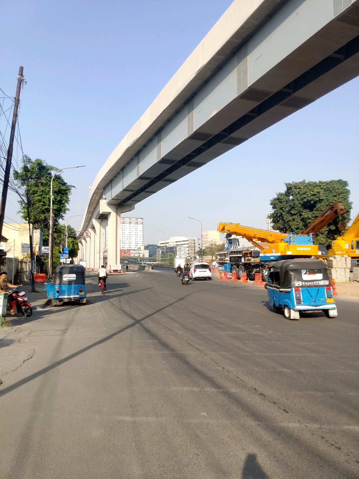 The image shows a wide, paved road in an urban setting, likely in East Jakarta, Indonesia, under a large, elevated concrete structure. The sky is clear and blue, indicating daylight. On the left side of the road, there are trees and utility poles with numerous wires. A man is seated on a red motorcycle parked by a pole, while another man is standing nearby. A blue three-wheeled vehicle, commonly known as a bajaj or bemo, is parked on the side of the road.
Further down the road, another blue bajaj is in motion, as is a white car. A person is riding a bicycle in the middle lane. On the right side of the road, construction equipment is visible, including a large yellow crane. Construction materials, such as stacked concrete blocks, are also present. The road itself has some wear and tear, with cracks and debris. Traffic cones are placed in the construction area. Several other vehicles, including motorcycles, are seen in the distance. The overall impression is of an active, somewhat congested urban environment undergoing infrastructure development.