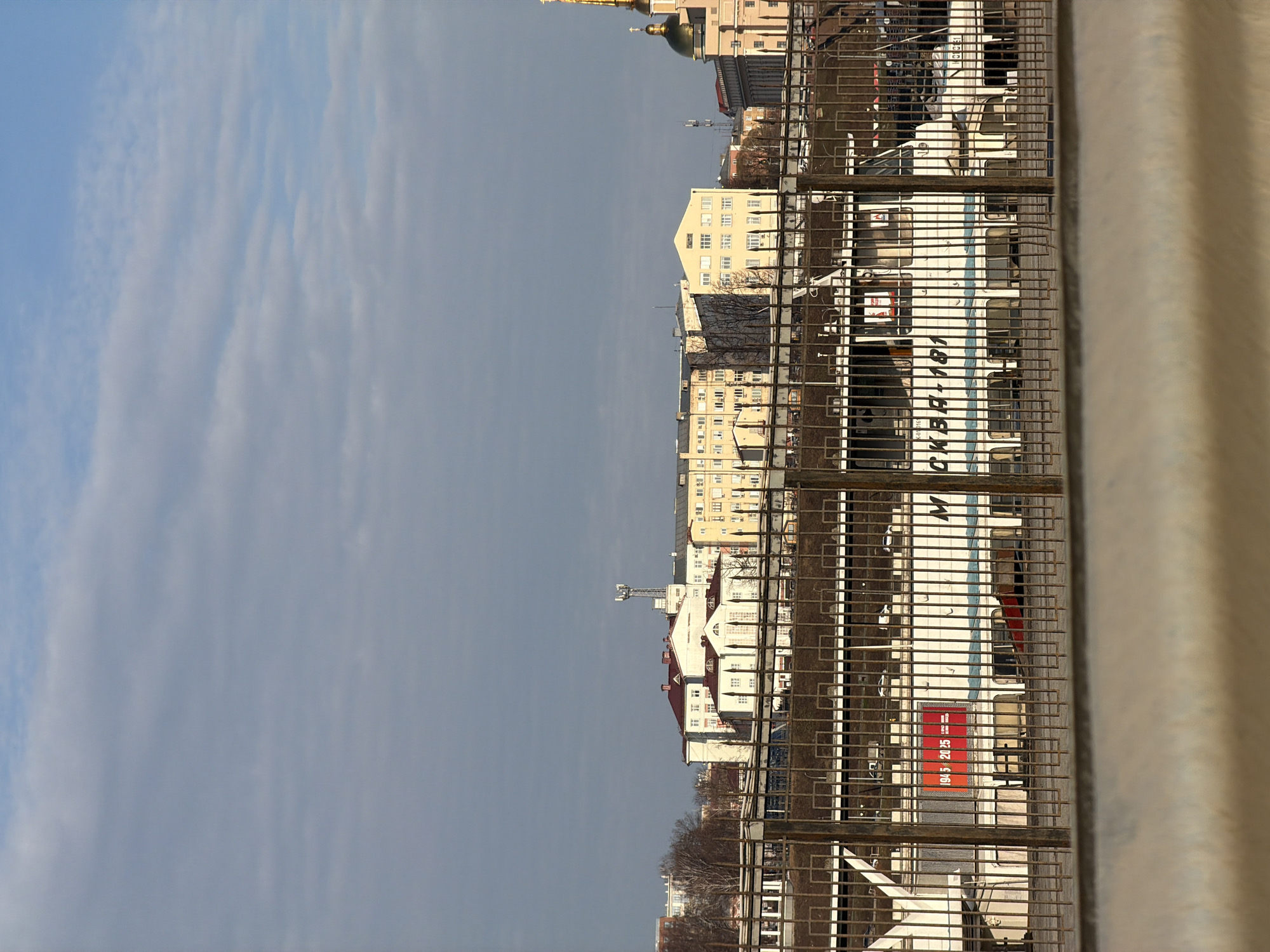 The image, taken from an elevated position behind a metal railing, shows an urban landscape in Izhevsk, Russia. Buildings of varying heights and architectural styles, including one with a golden dome, are visible in the background. A large, white building with blue and red accents is prominent, and the text "ОКВАН 181" is visible. A banner with the numbers "1945-2025" is visible. The sky above is partly cloudy.