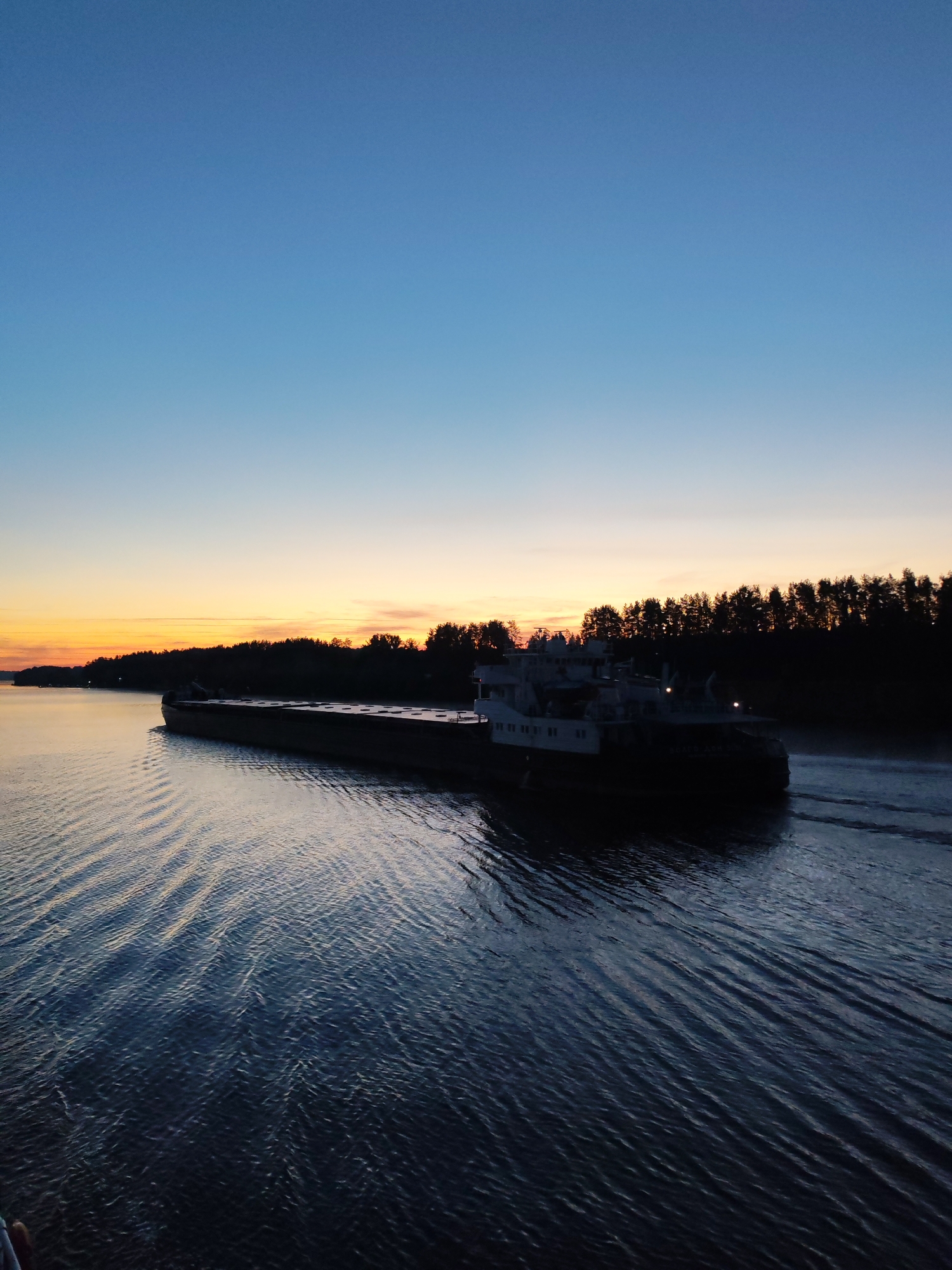 The image displays a twilight scene over a calm body of water in the city of Unknown, Unknown. The sky presents a gradient, transitioning from a deep blue at the top of the frame to a lighter blue mid-frame, and then to a band of bright orange and yellow along the horizon. A dense line of silhouetted trees, predominantly conifers, is visible along the distant bank, positioned against the brighter part of the horizon. A large, dark-colored cargo vessel or barge, featuring a multi-deck superstructure at its stern, is positioned in the midground, moving from left to right. The vessel casts a dark silhouette against the sky, with small internal lights visible within its superstructure. Distinct parallel wakes emanate from the stern of the vessel, extending across the dark blue water. The water surface exhibits numerous ripples and reflections of the sky's varied hues. No individuals are discernible within the frame.