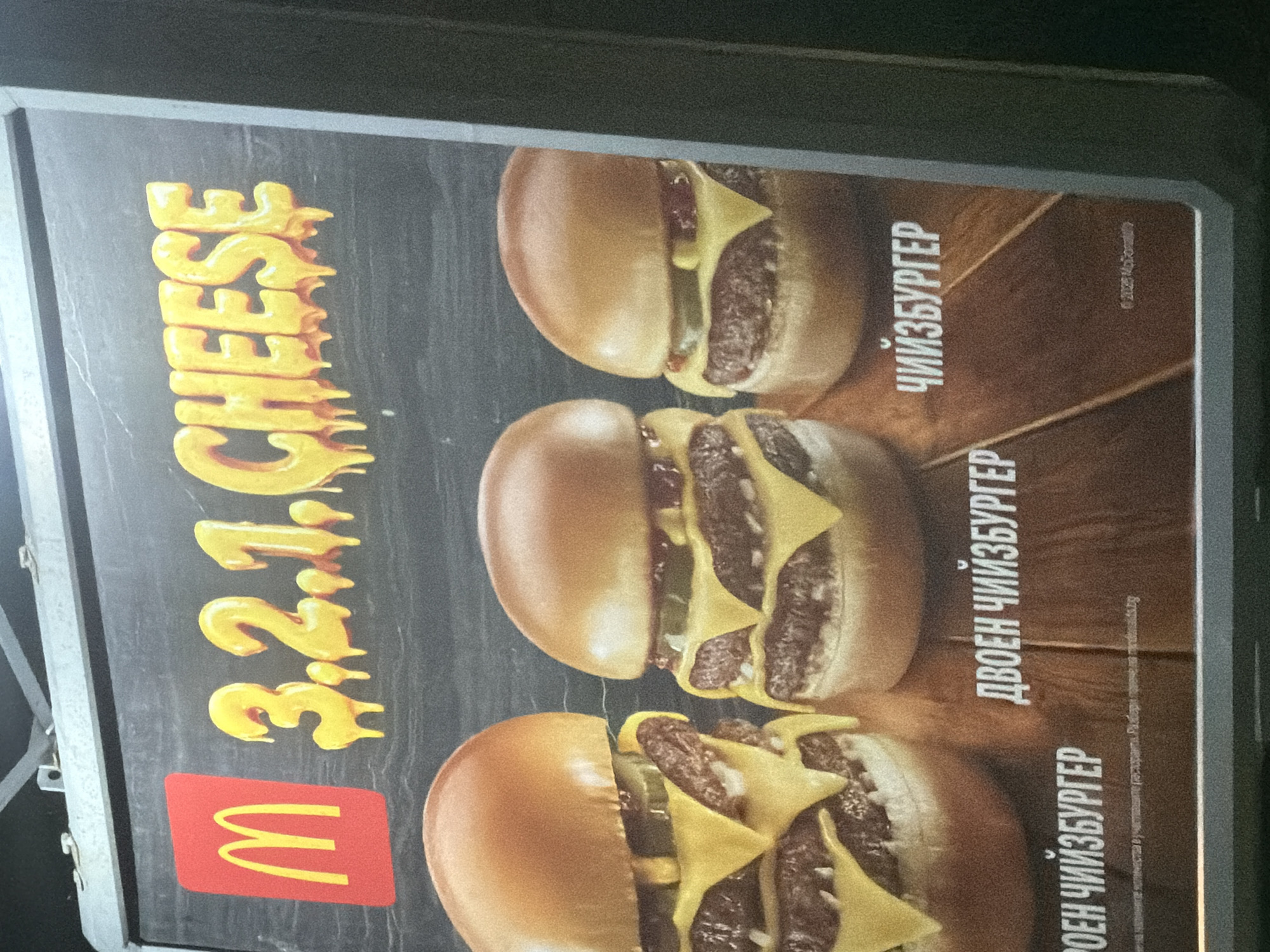 An outdoor McDonald's advertisement is displayed on a light board, featuring three cheeseburgers against a dark background with a subtle wooden texture. The image shows the advertisement rotated clockwise.
The main subjects are three cheeseburgers, presented vertically. From top to bottom, there's a single-patty cheeseburger, a double-patty cheeseburger, and a triple-patty cheeseburger, each with melted cheese and pickles visible between the patties and buns.
To the left of the cheeseburgers, the text "3.2.1. CHEESE" is prominently displayed in a bold, yellow font with a melted cheese effect, indicating a sequence of cheese burgers. Below this text, the iconic red and yellow McDonald's "M" logo is visible.
To the right of the cheeseburgers, their names are written in Cyrillic, likely Bulgarian, which aligns with the provided location context of Burgas, Bulgaria. From top to bottom, the text reads:
* "ЧИЙЗБУРГЕР" (Cheeseburger)
* "ДВОЕН ЧИЙЗБУРГЕР" (Double Cheeseburger)
* "ТРОЕН ЧИЙЗБУРГЕР" (Triple Cheeseburger - inferred from the visual despite partial OCR results)
In the bottom right corner, smaller text includes a copyright notice: "© 2022 McDonald's" and a disclaimer in Bulgarian, which translates to: "All prices and quantities are approximate for participating restaurants. Find out more at mcdonalds.bg".
The scene appears to be at night or in low light, given the illuminated nature of the display and the dark surroundings of the advertising board frame. No people or specific activity are depicted beyond the advertisement itself.