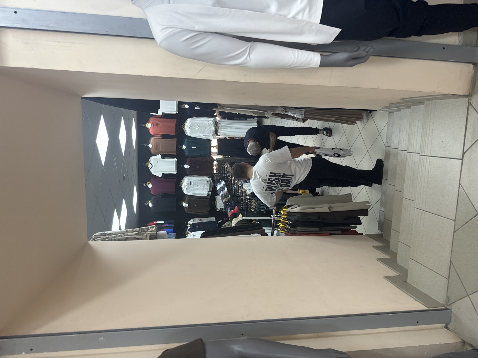 The image shows the interior of a clothing store in Saint Petersburg, Russia. The store appears to be a multi-level establishment, with a staircase in the foreground leading down to the main shopping area. The walls are painted a light peach color, and the ceiling is a drop ceiling with fluorescent lighting.
In the main shopping area, racks of clothing are displayed. There are men's t-shirts and shirts hung on display. Several people are visible in the store. In the foreground, on the stairs, a person wearing a white t-shirt with "PUSH SOME" printed on it is seen looking down. Another person, wearing a dark t-shirt and dark pants, is bent over, possibly examining clothing on a lower rack. On the right side of the image, a mannequin wearing a white long-sleeved shirt is visible.
The overall impression is a casual retail environment, likely a small to medium-sized clothing boutique.