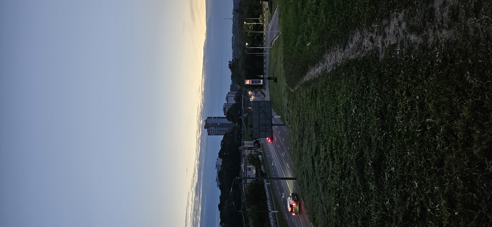 The image depicts a twilight scene of a road and cityscape in Vitsebsk, Belarus, likely near the intersection of Moskovsky Prospekt and Vulitsa Maskovkaja, given the presence of a LUKOIL gas station. In the foreground, a grassy hill slopes down towards a paved road where two vehicles are visible. The vehicle on the left is a dark-colored car with its red taillights illuminated, driving away from the viewer. Further down the road, another vehicle with illuminated red taillights is also visible, suggesting traffic is moving. A pedestrian pathway made of dirt and gravel is visible in the grassy area, leading towards the road.
In the mid-ground, several buildings are situated along the road, including apartment buildings and a LUKOIL gas station with illuminated signage. Trees and streetlights line the road, and a large, empty billboard is positioned on the right side of the road.
The background features a line of trees separating the road from a cluster of taller apartment buildings. The sky is a gradient of pale blue and grey, with a band of darker clouds near the horizon where the sun has recently set, casting a soft, diffused light. The overall atmosphere is calm and quiet, indicative of the transition from day to night.